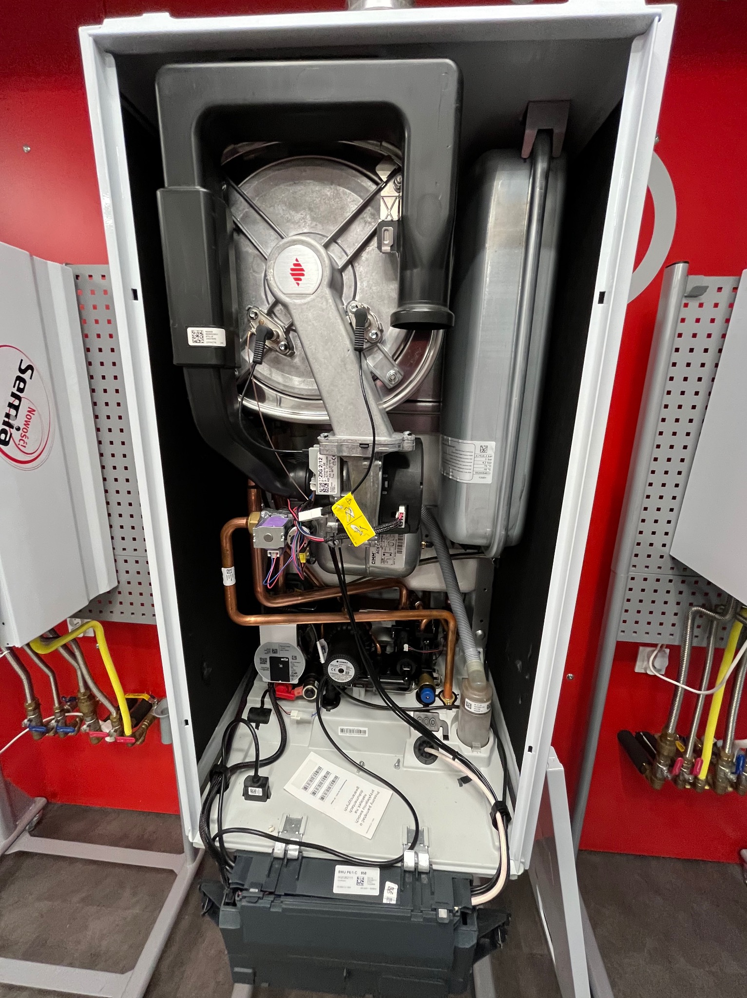 Wnętrze kotła gazowego z widocznymi komponentami: palnikiem, wymiennikiem ciepła, rurami miedzianymi i elektroniką. Widoczne oznaczenia producenta i etykiety informacyjne.