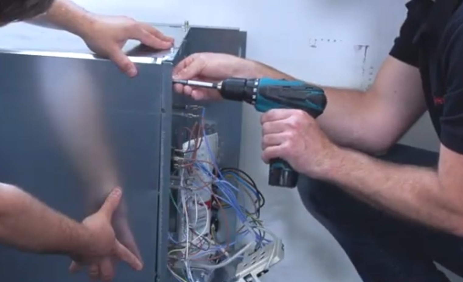 Monterażysta skręca obudowę kotła gazowego w pomieszczeniu. Widoczne przewody i elementy wewnętrzne kotła. Używa wkrętarki akumulatorowej.