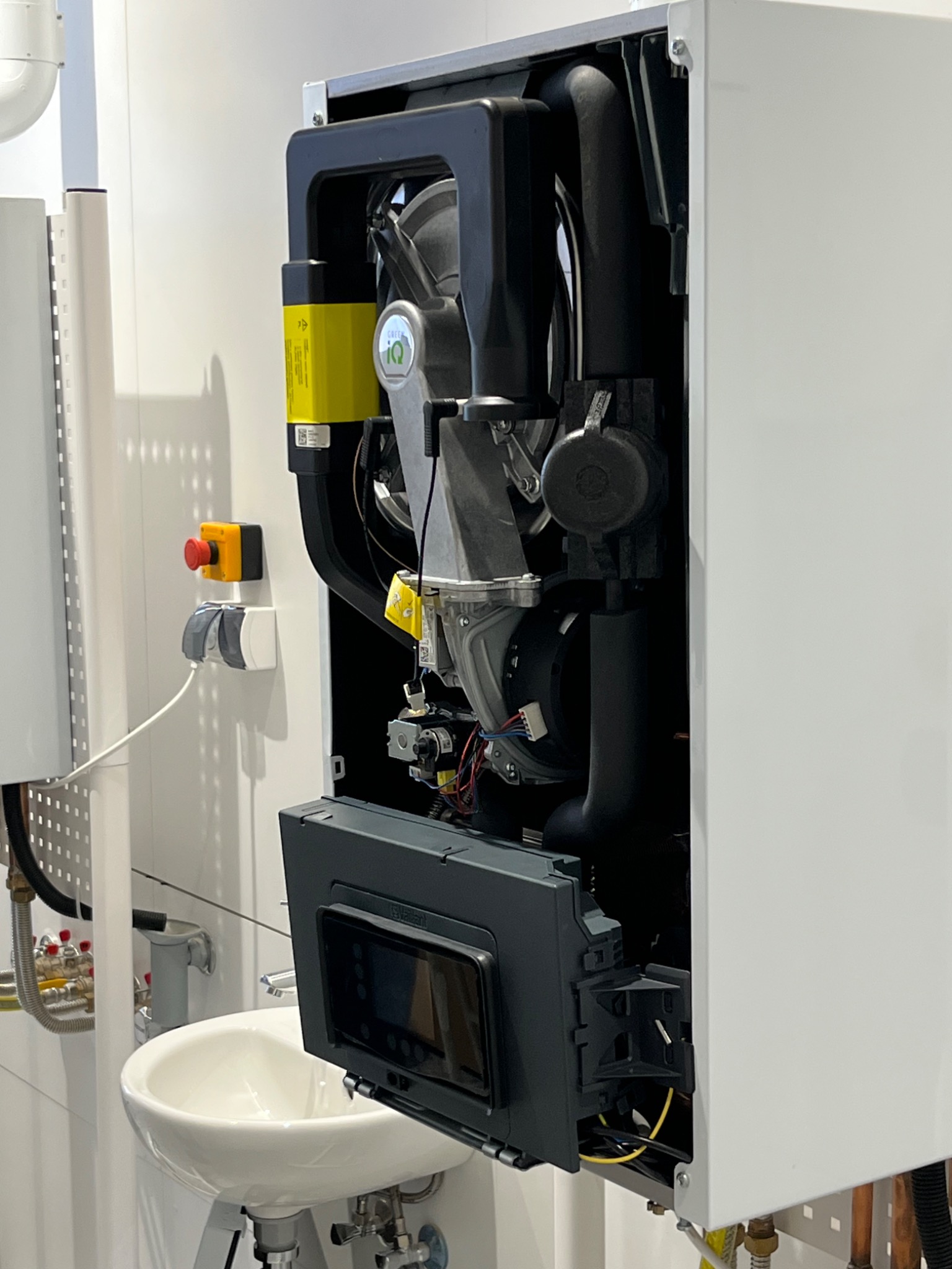 Otwarty, nowoczesny piec gazowy z widocznymi elementami wewnętrznymi, zamontowany na białej ścianie w pomieszczeniu z umywalką. Widoczne rury i instalacja.