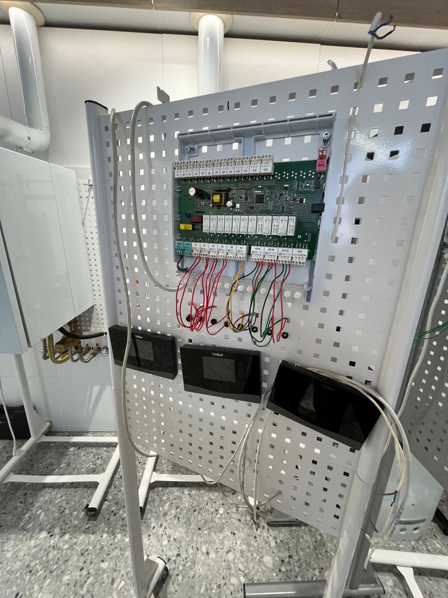 Panel demonstracyjny Vaillant z podłączonymi przewodami i sterownikami, umieszczony na perforowanej, białej konstrukcji. Widoczny fragment kotła gazowego. Prezentacja instalacji grzewczej.