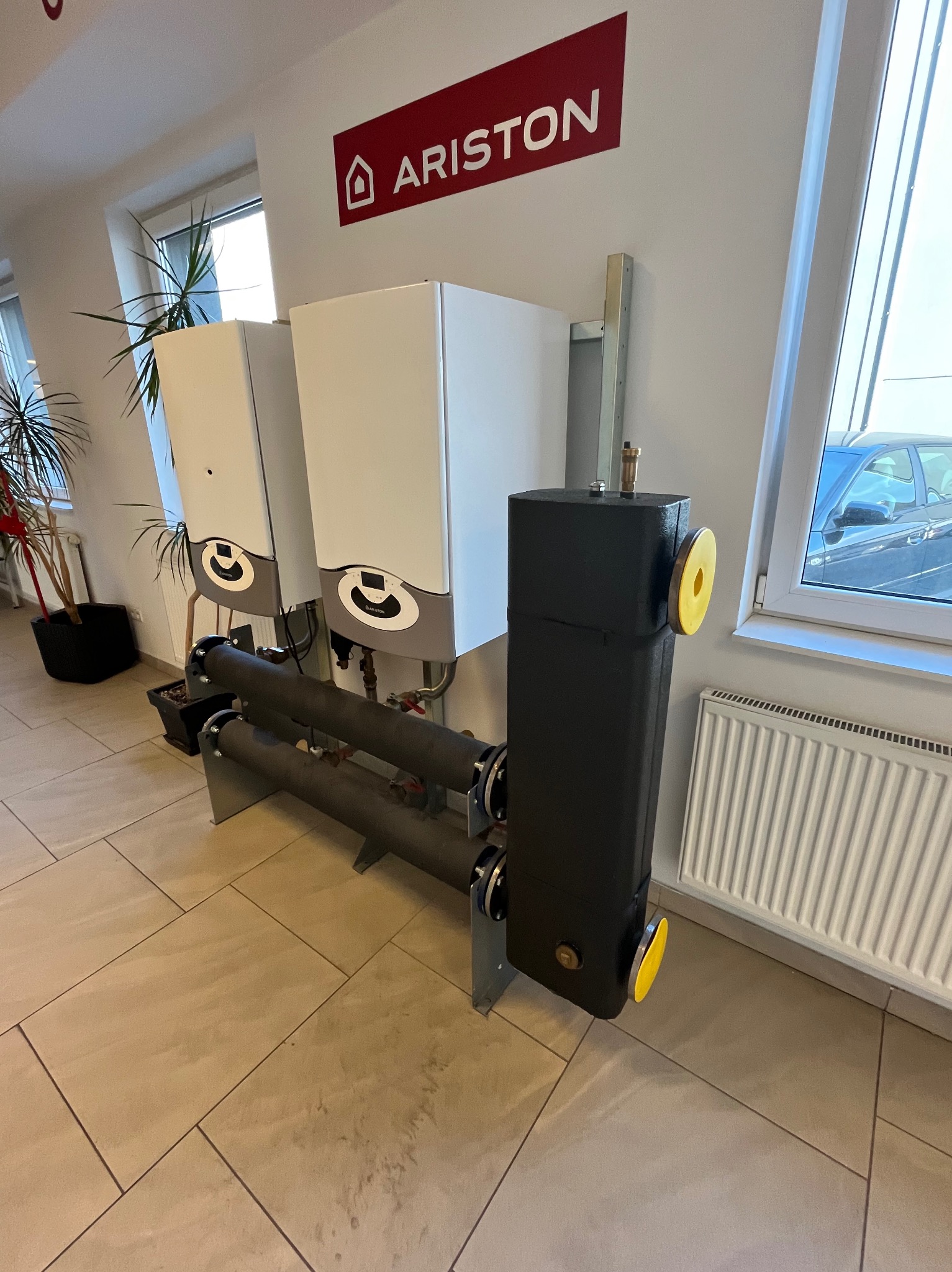 Dwa białe piece gazowe Ariston z czarnym zasobnikiem ciepłej wody, połączone rurami. Widoczny grzejnik i roślina doniczkowa. Jasne wnętrze, płytki podłogowe.
