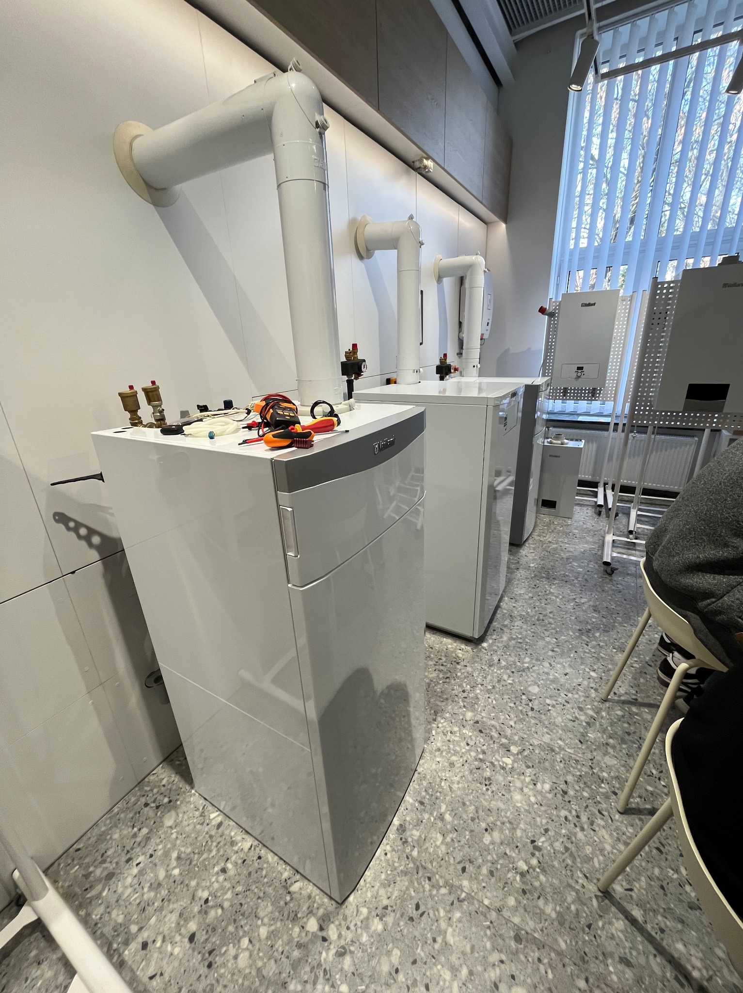 Białe, wiszące piece gazowe Vaillant z podłączonymi rurami, narzędzia na górze. Widok na okno z żaluzjami. Szary wzór na podłodze. Częściowo widoczna osoba.