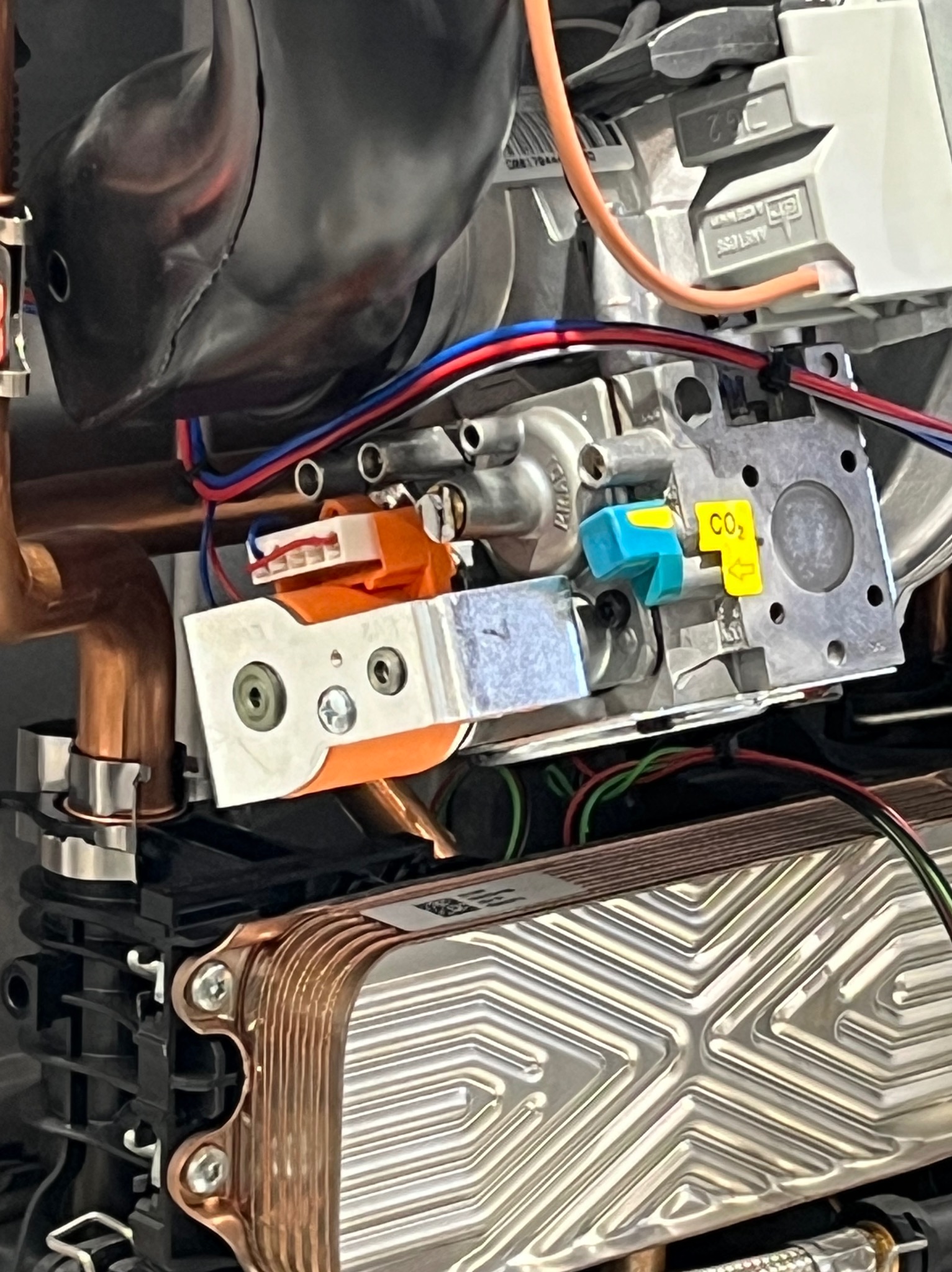 Wnętrze kotła gazowego z widocznymi elementami: wymiennik ciepła, zawory, przewody elektryczne i rury miedziane. Detaliczny widok na podzespoły urządzenia grzewczego.