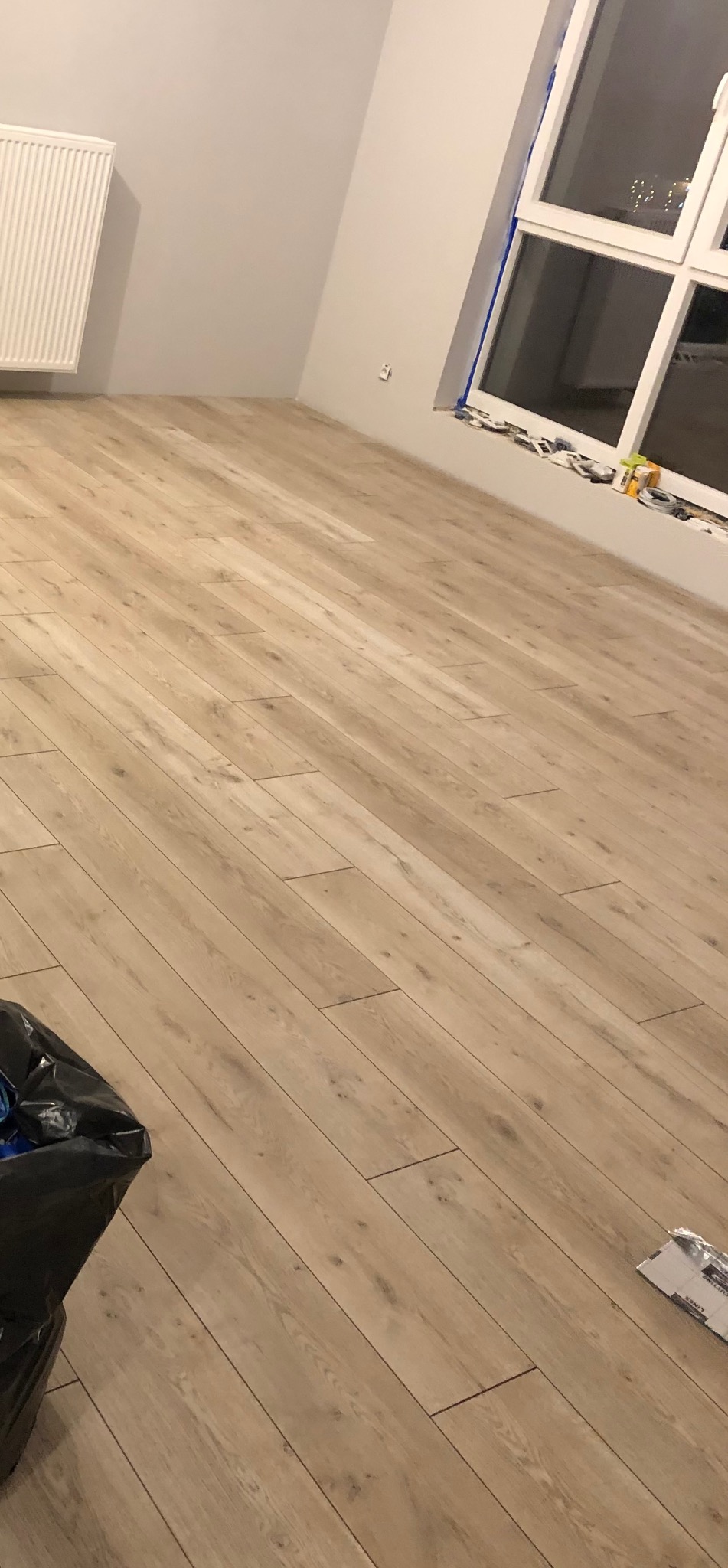 Jasne panele podłogowe ułożone w pokoju w Nowym Dworze Mazowieckim. Widoczny fragment okna, grzejnik i śmieci. Podłoga o wzorze drewna.