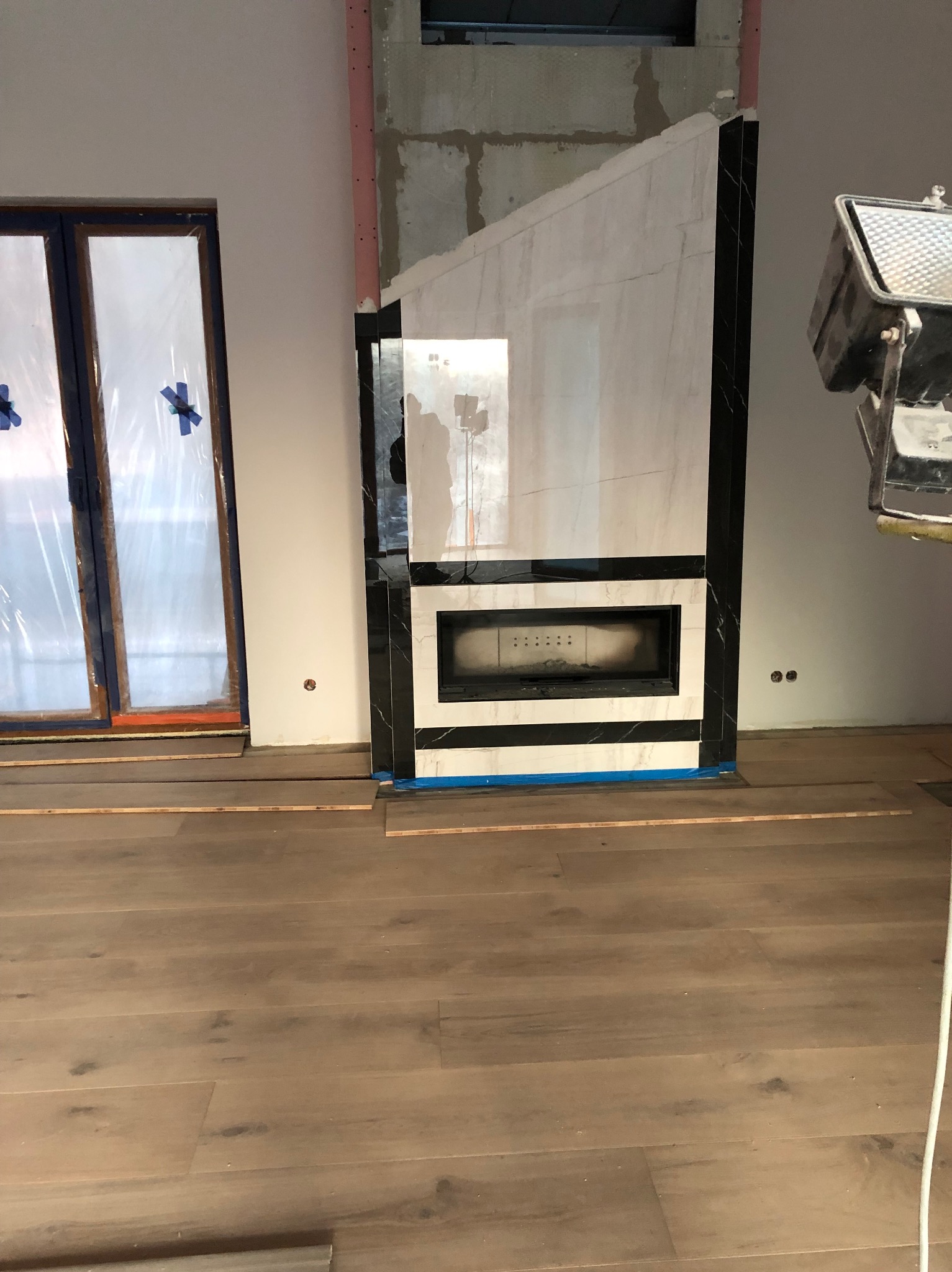 Wnętrze w trakcie prac wykończeniowych: podłoga z paneli, marmurowy kominek, zabezpieczone folią drzwi balkonowe. Widoczne elementy oświetlenia budowlanego.