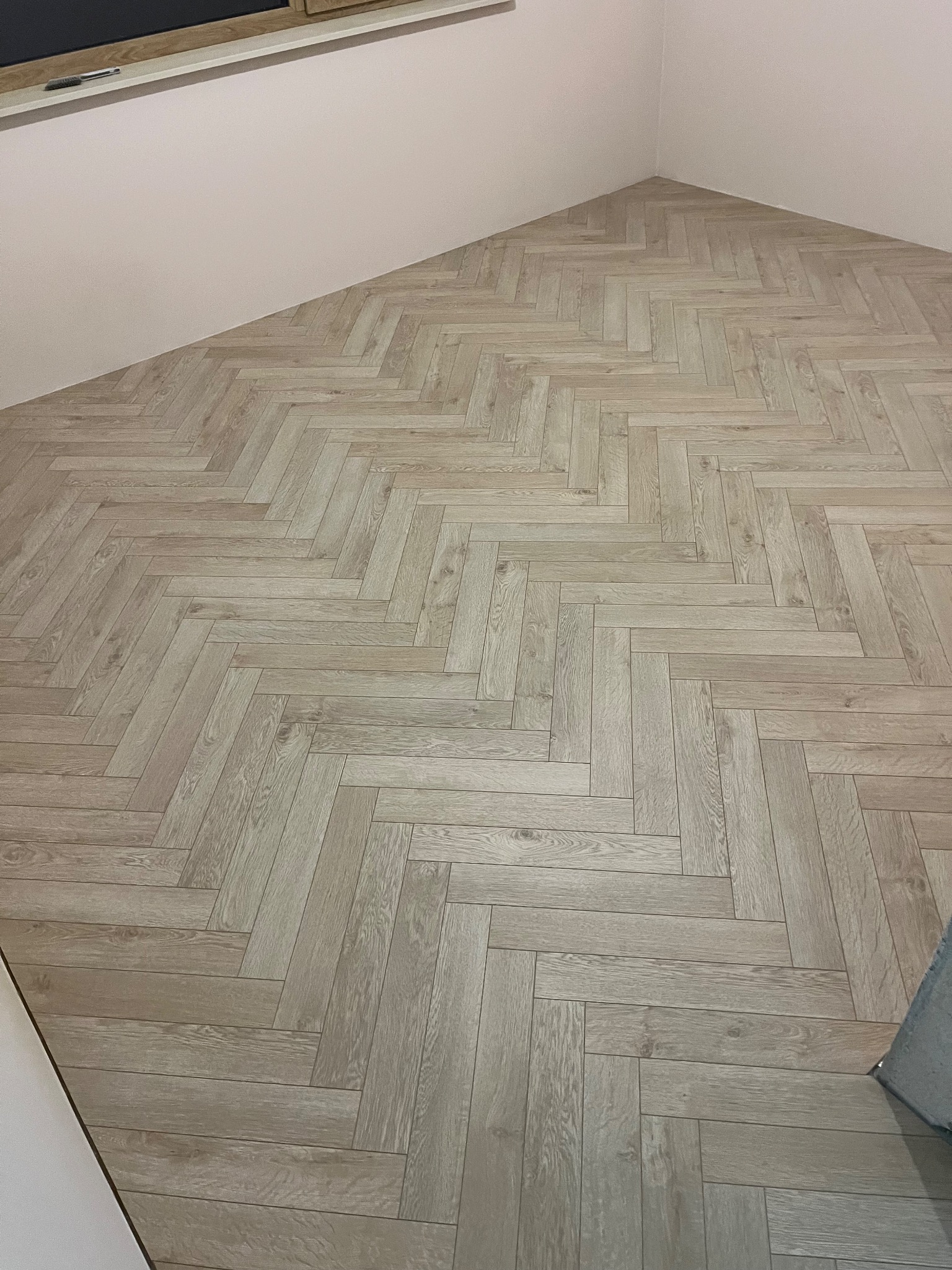 Jasne panele ułożone w jodełkę na podłodze w rogu pokoju. Widoczne ściany w jasnym odcieniu. Ujęcie z góry, prezentujące wzór i kolorystykę podłogi.