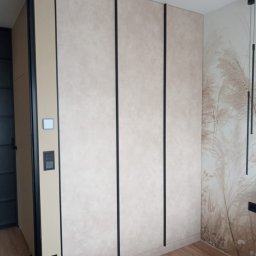 KAROLINA SOŁOBODOWSKA - Beżowa szafa wnękowa z czarnymi, pionowymi akcentami, dopasowana do wnętrza sypialni z tapetą w motywy roślinne i drewnianą podłogą.