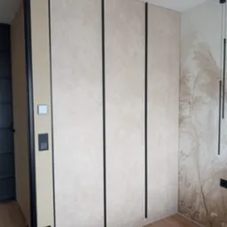 Beżowa szafa wnękowa z czarnymi, pionowymi akcentami, dopasowana do wnętrza sypialni z tapetą w motywy roślinne i drewnianą podłogą.