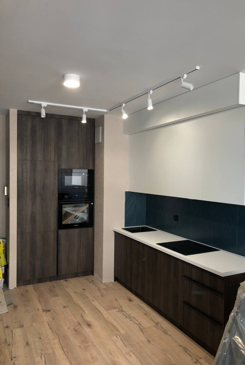 Nowoczesna kuchnia: ciemne, drewnopodobne szafki z białym blatem i niebieską mozaiką. Oświetlenie szynowe podkreśla detale.