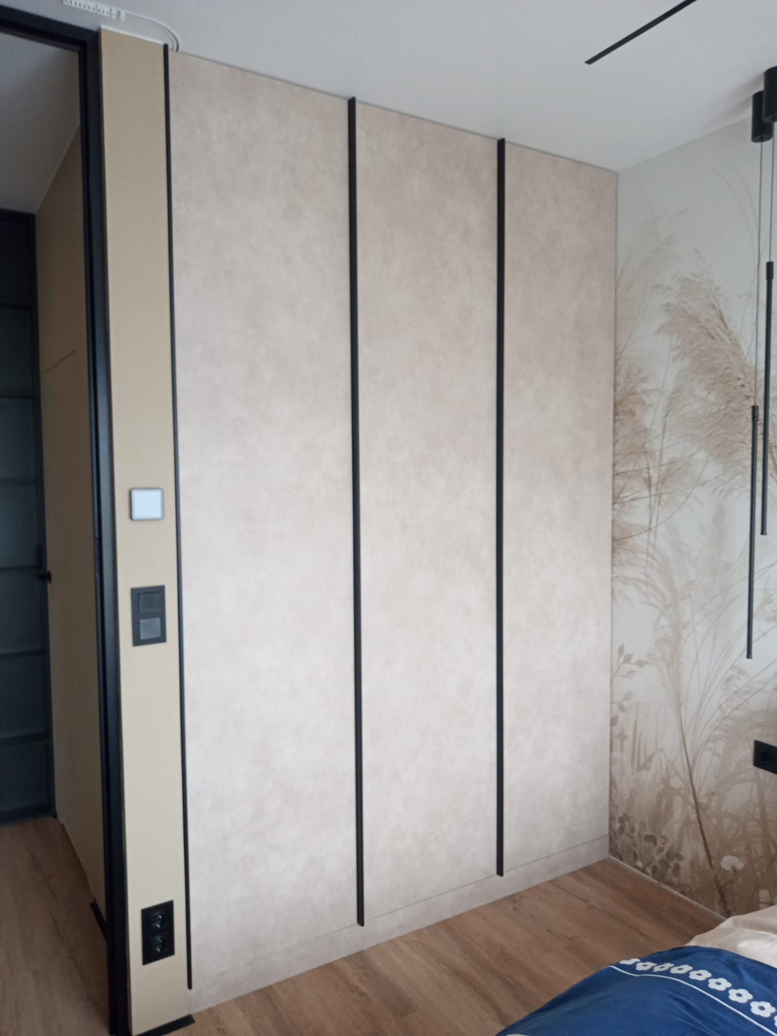 Beżowa szafa wnękowa z czarnymi, pionowymi akcentami, dopasowana do wnętrza sypialni z tapetą w motywy roślinne i drewnianą podłogą.