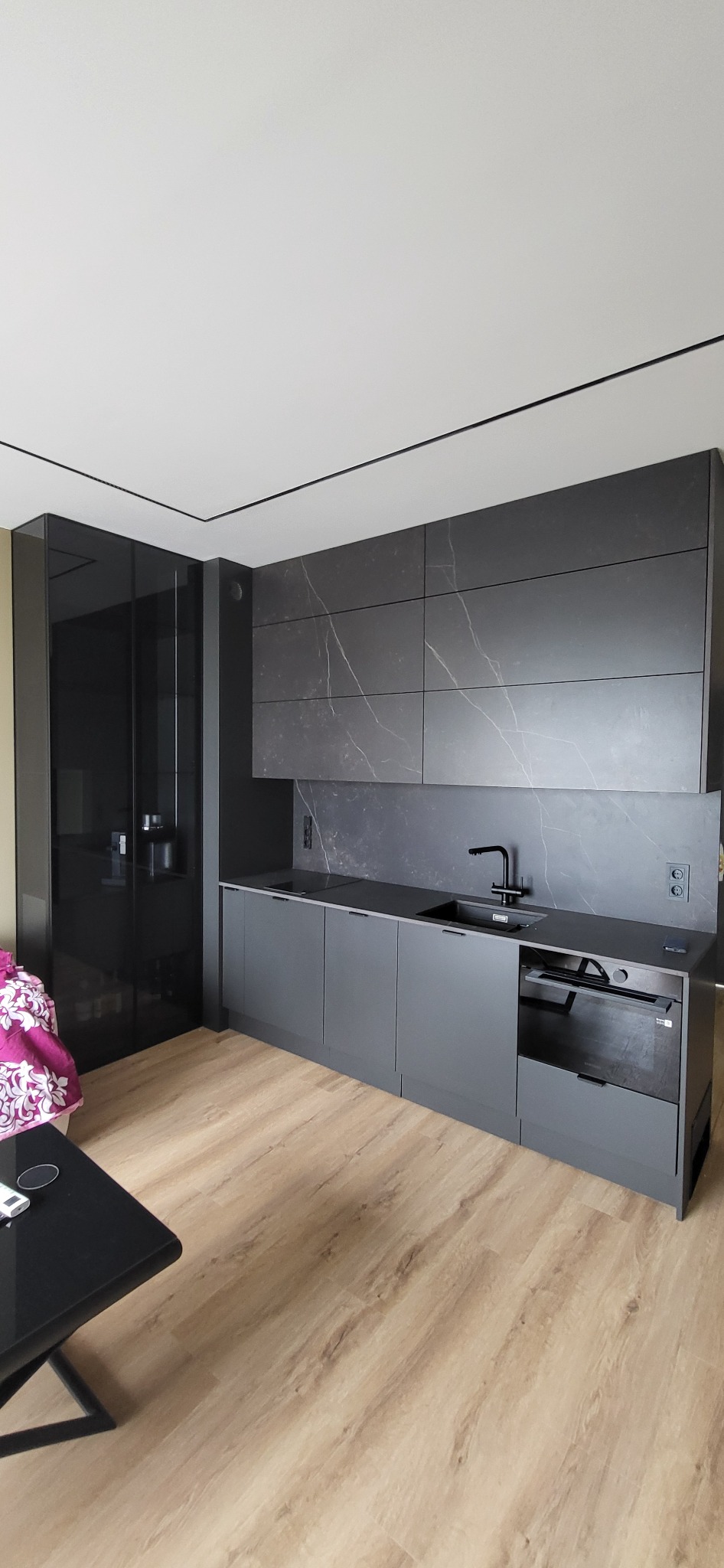 Nowoczesna, szara kuchnia z czarnymi akcentami: armatura, fronty szafek i blat. Płytki imitujące marmur na ścianie. Widoczna zabudowa piekarnika i szafa.