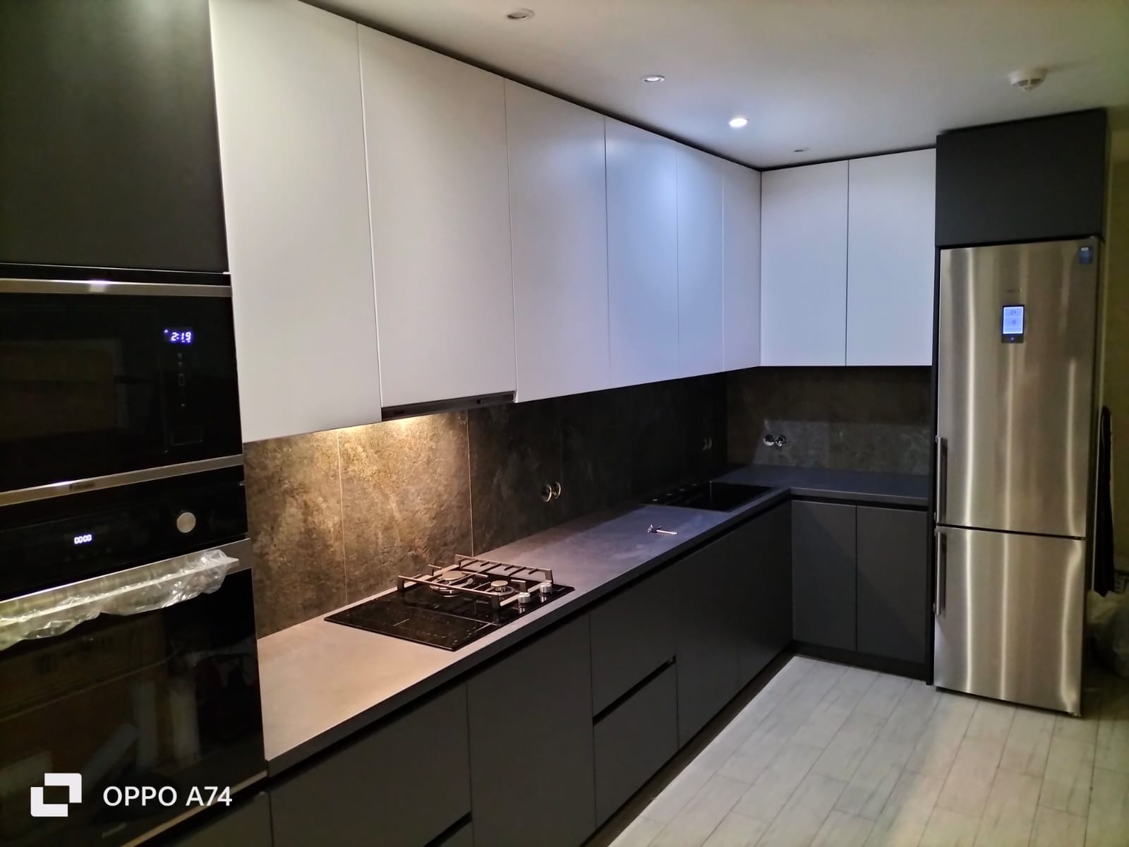 Nowoczesna kuchnia z szarymi szafkami dolnymi i białymi górnymi, wykończona kamiennym blatem i stalową lodówką. Minimalistyczny design z oświetleniem punktowym.