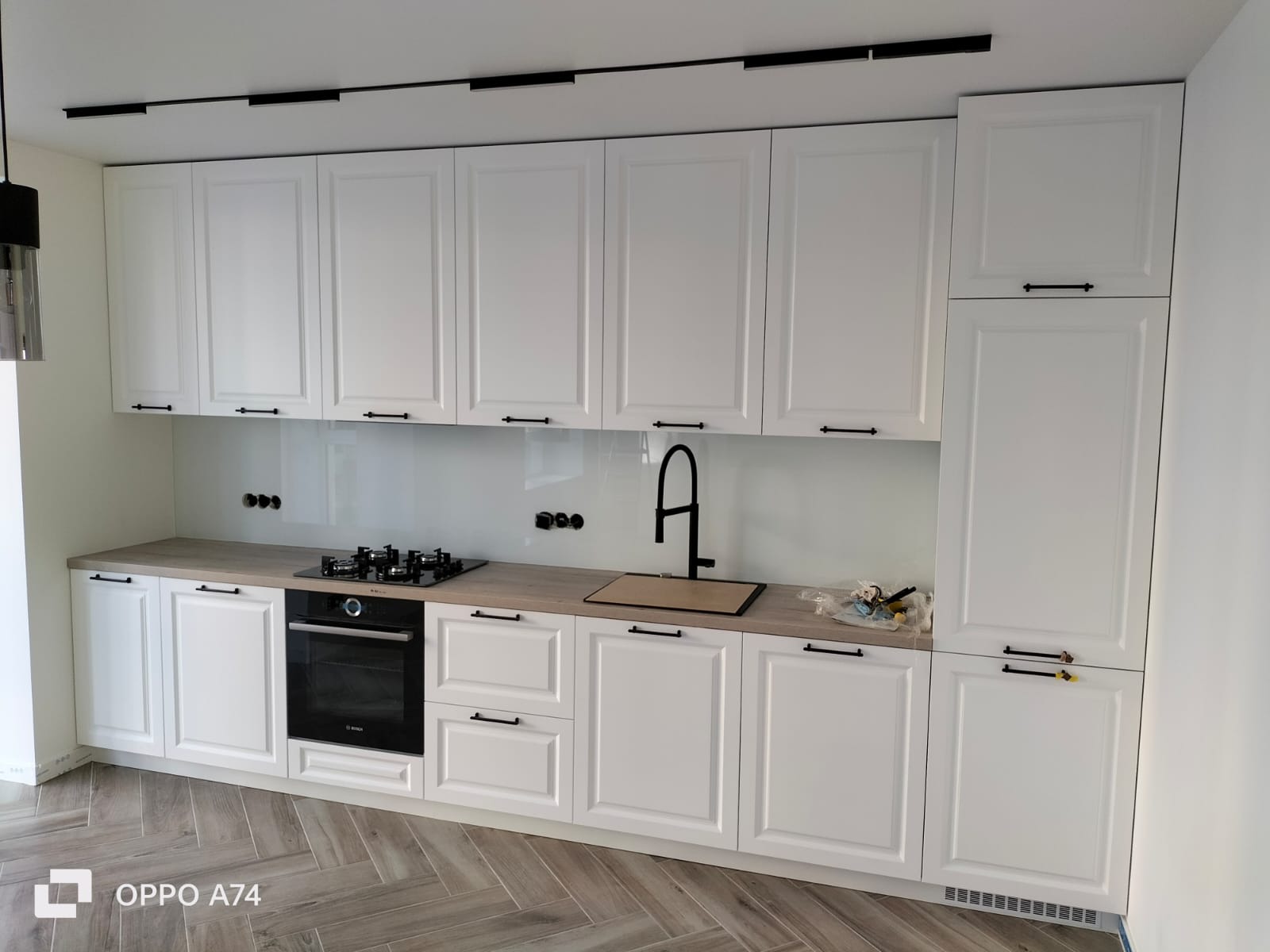 Biała kuchnia na wymiar z czarnymi akcentami: bateria, uchwyty, płyta grzewcza. Jasny blat i backsplash. Podłoga w jodełkę. Nowoczesny design.