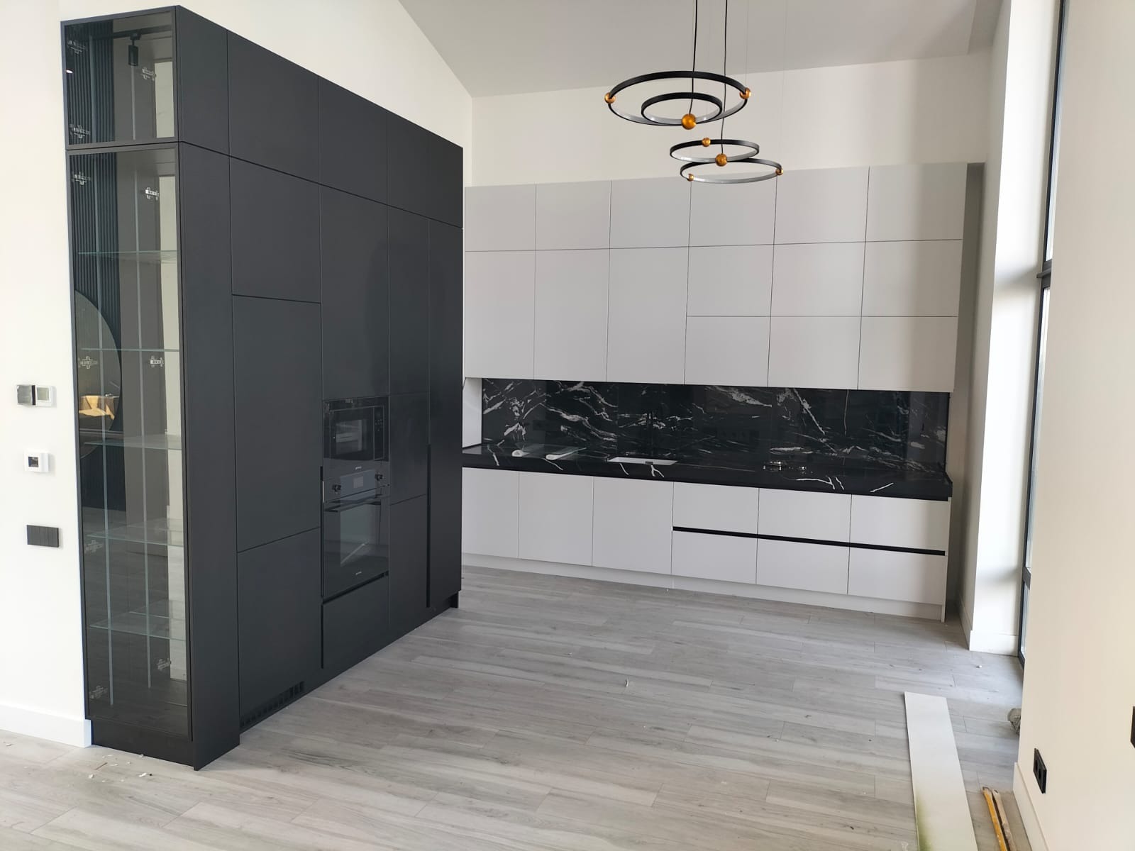 Nowoczesna kuchnia z zabudową w odcieniach szarości i bieli, z czarnym blatem imitującym marmur. Wiszące lampy w kształcie okręgów dodają elegancji.