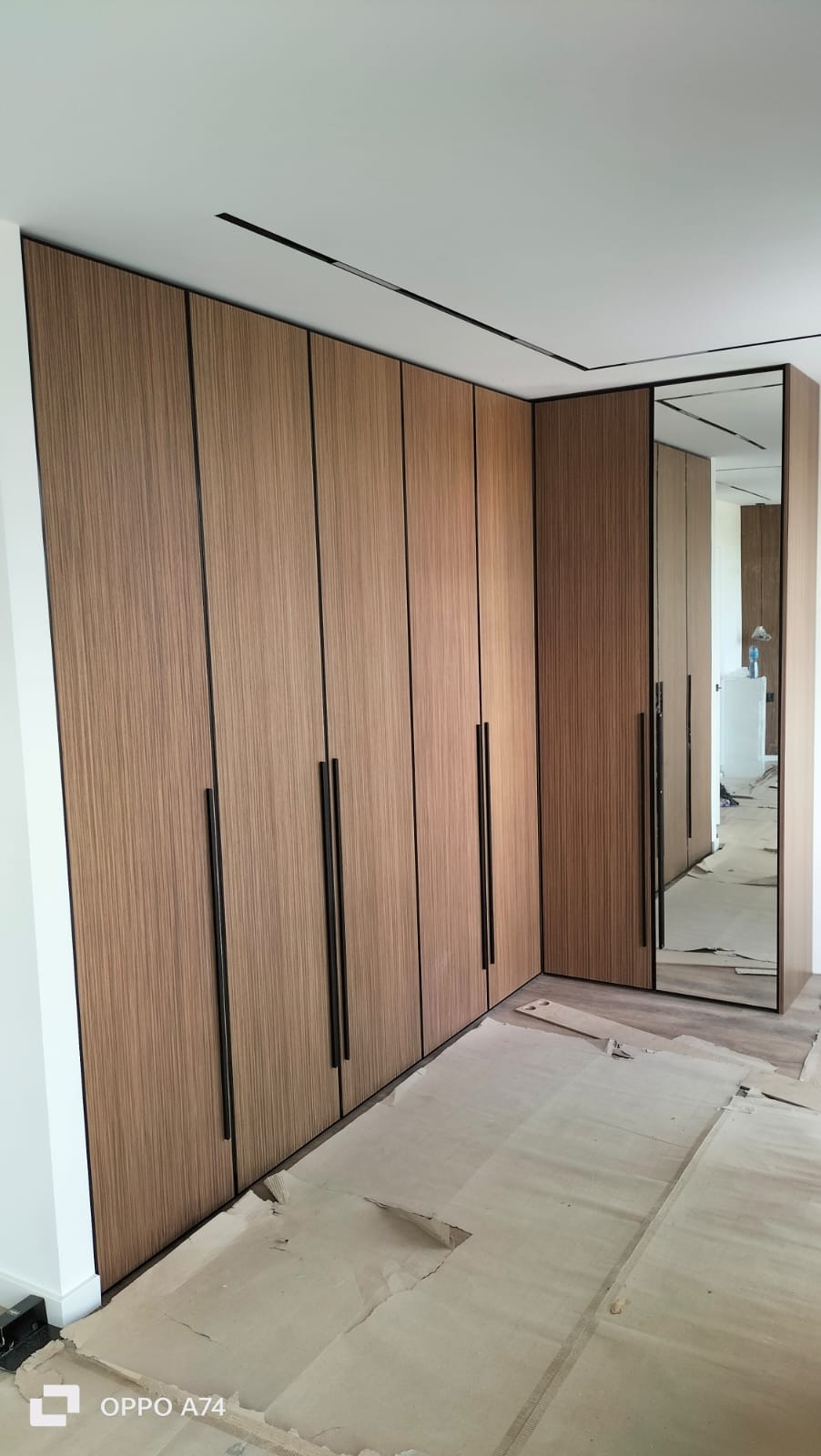 Szafa wnękowa z lustrem i drewnianymi frontami, czarne, długie uchwyty, widok na narożnik szafy, podłoga zabezpieczona kartonem, minimalistyczny design.