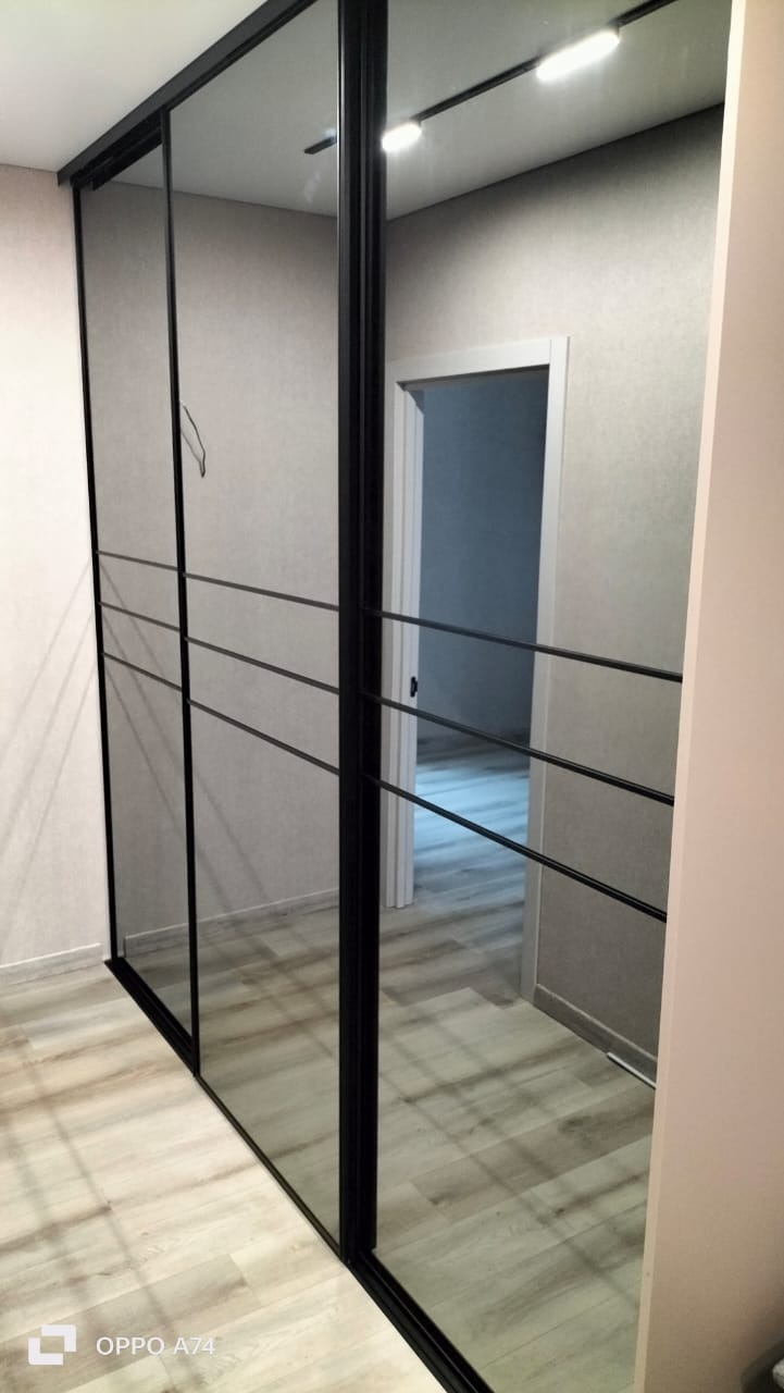 Nowoczesna szafa wnękowa z czarnymi ramami i lustrzanymi drzwiami przesuwnymi. Widoczne oświetlenie sufitowe i fragment wnętrza pokoju. Minimalistyczny design.