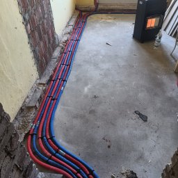 Hydraullica - Kamil Dziuba - Równolegle ułożone, czerwono-niebieskie rury instalacji grzewczej w bruzdach podłogi, z widocznym piecem przenośnym i fragmentem ściany z cegły.