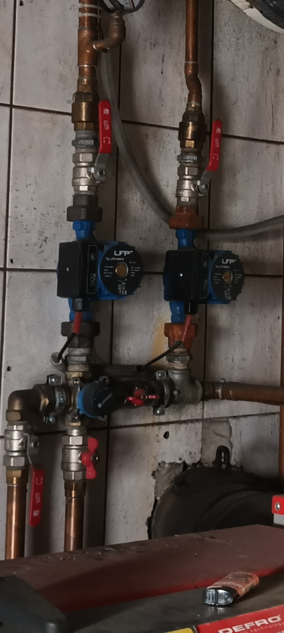 Złożona instalacja grzewcza z miedzianymi rurami, zaworami z czerwonymi uchwytami i dwoma pompami obiegowymi LFP, na tle kafelkowanej ściany.