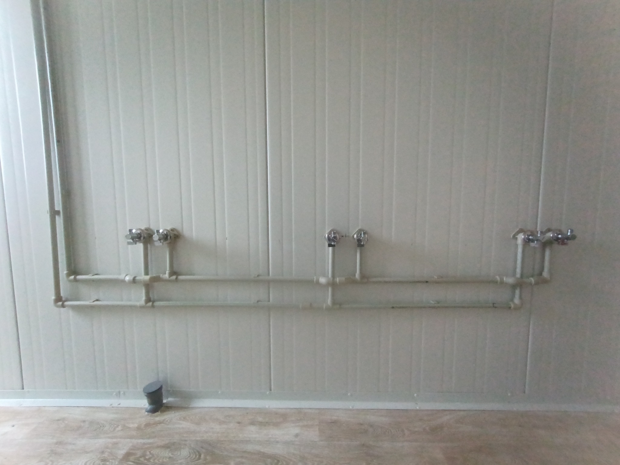 Szare rury instalacji wodnej z zaworami, zamontowane na panelowej ścianie. Na podłodze widoczny fragment odpływu. Ujęcie z bliska, prezentujące detale instalacji.