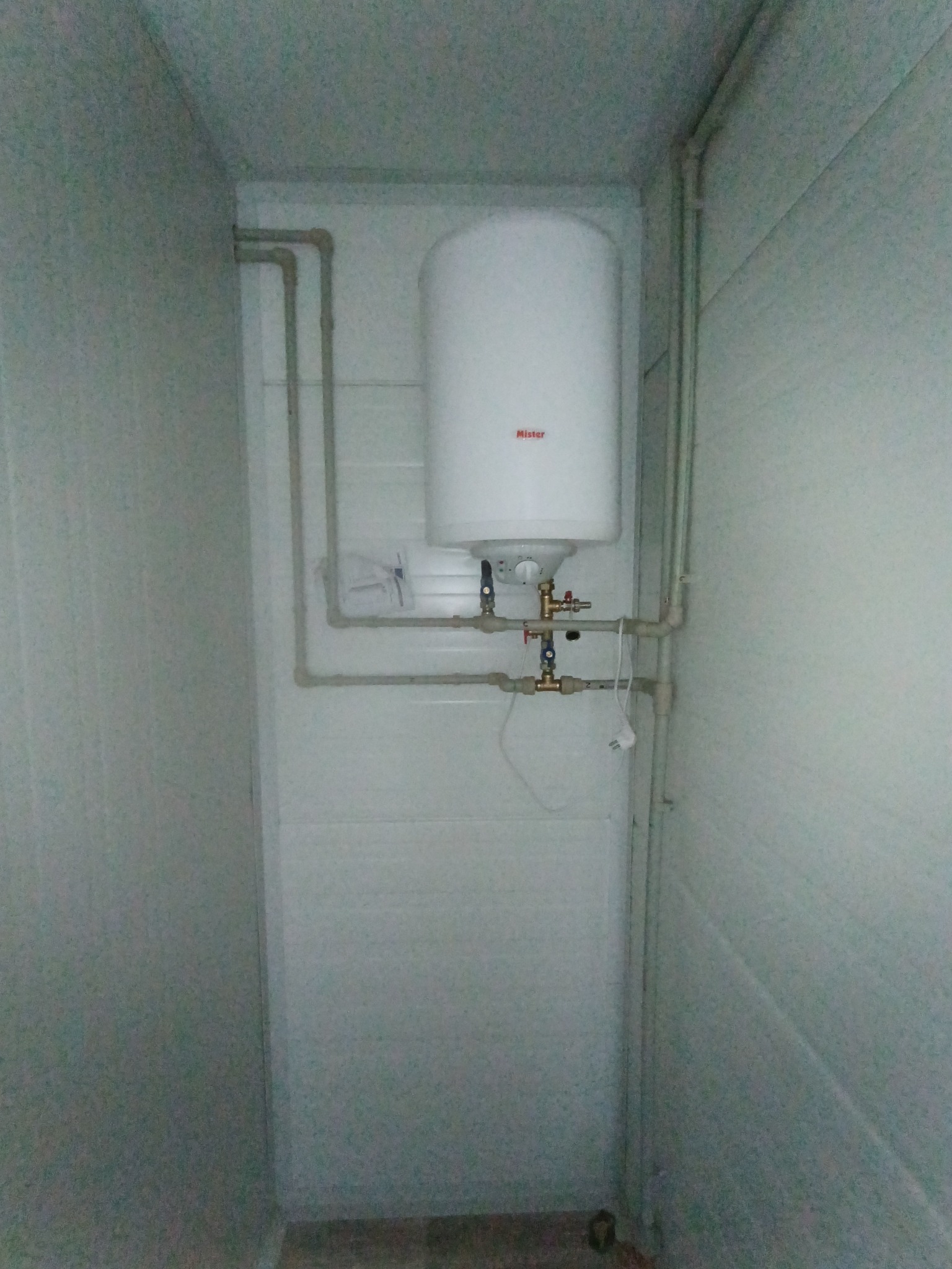Zamknięta przestrzeń z zamontowanym bojlerem Mister i instalacją hydrauliczną z zaworami. Widoczne rury i przyłącza. Ściany wykończone panelami.