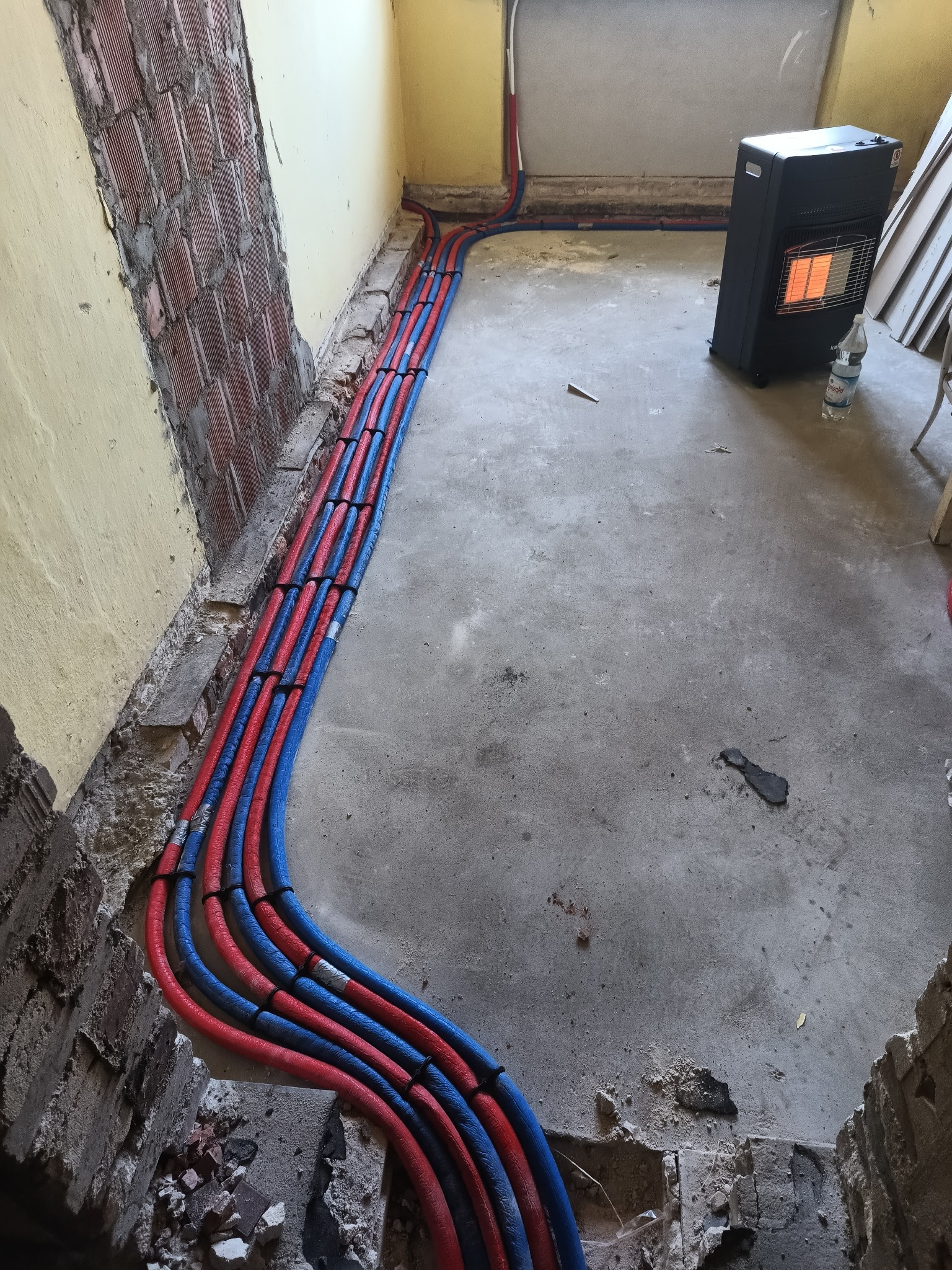 Równolegle ułożone, czerwono-niebieskie rury instalacji grzewczej w bruzdach podłogi, z widocznym piecem przenośnym i fragmentem ściany z cegły.