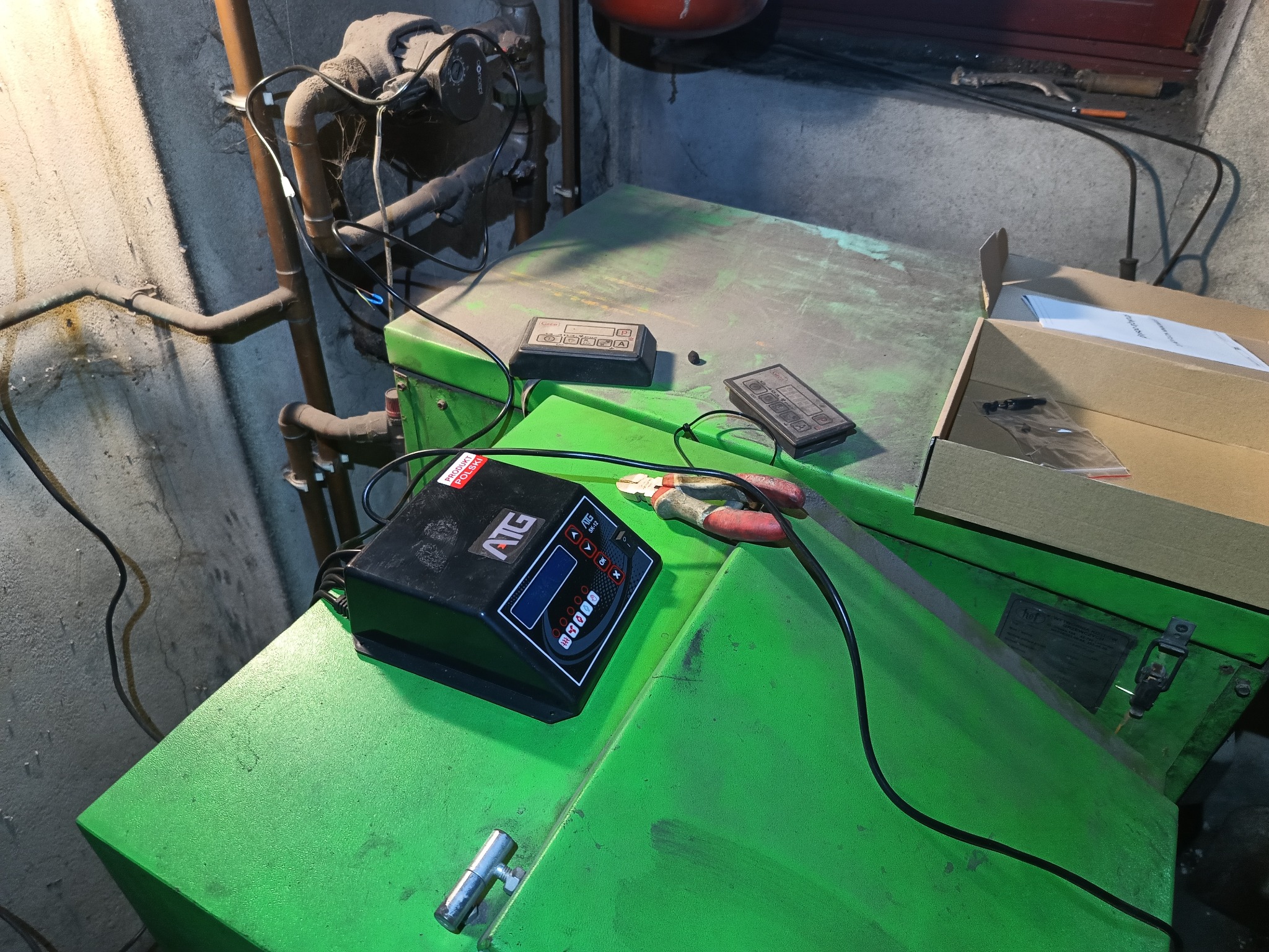 Zielony piec CO z podłączonym testerem ATG SK-12, widoczne rury instalacji, narzędzia i karton. Ujęcie z góry, oświetlenie mieszane.