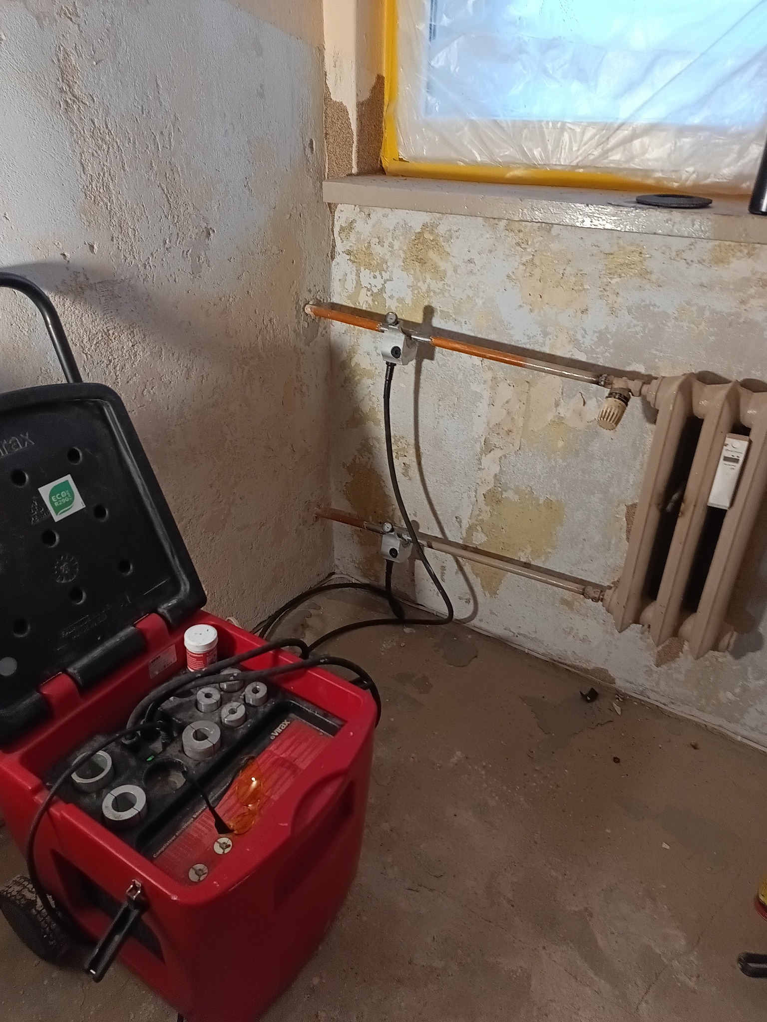 Podłączenie grzejnika żeliwnego do instalacji centralnego ogrzewania. Widoczna zgrzewarka do rur i fragment okna zabezpieczonego folią malarską.