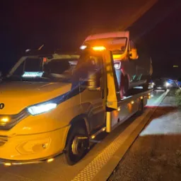 Żółta laweta Iveco z włączonymi światłami ostrzegawczymi, transportująca busa nocą na autostradzie. Widoczne światła innych pojazdów.