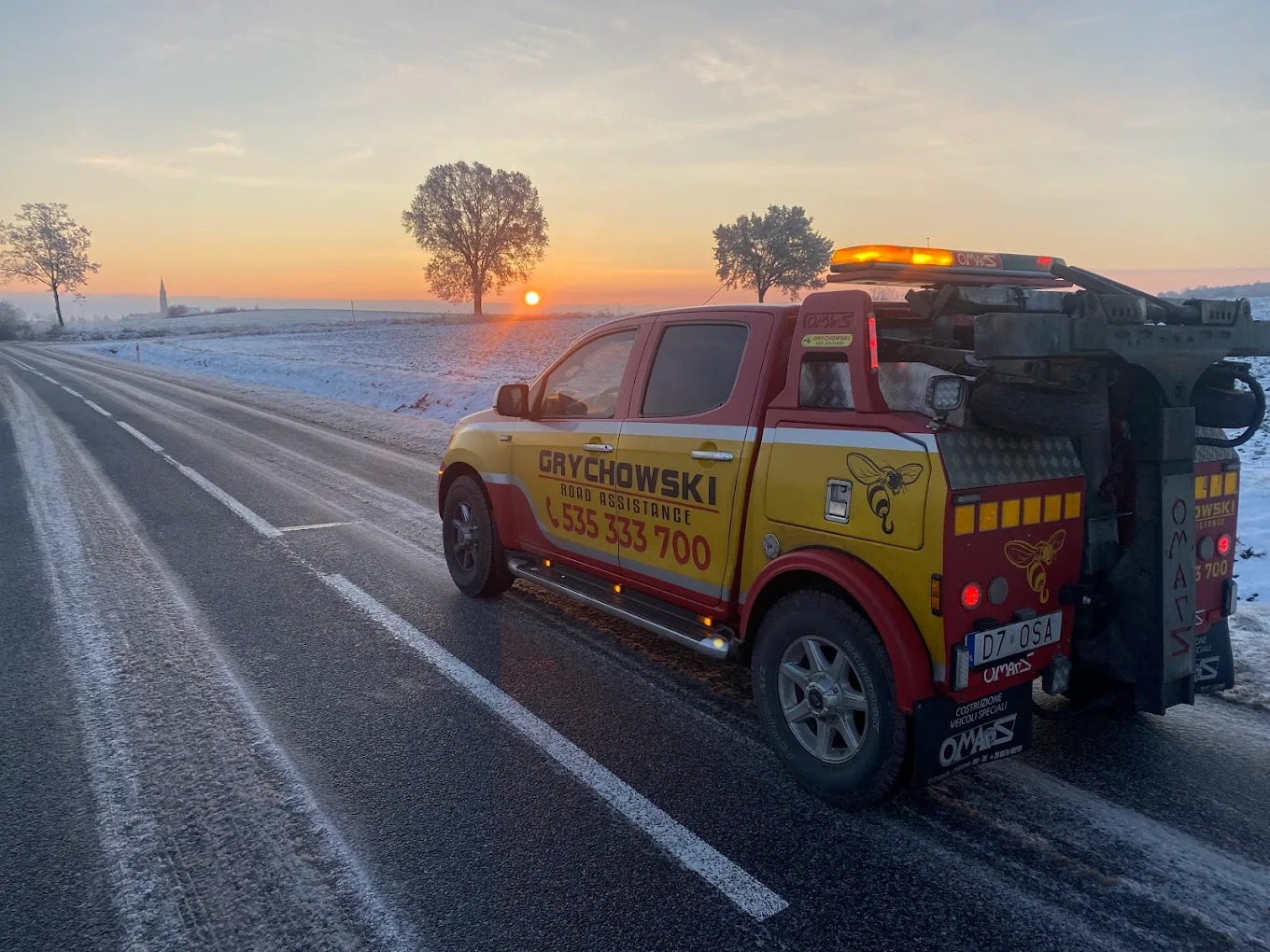 Żółto-czerwona laweta Grychowski Road Assistance na ośnieżonej drodze o świcie, z widocznym logo pszczoły i numerem telefonu 535 333 700.