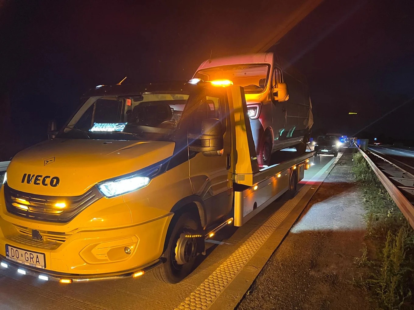 Żółta laweta Iveco z włączonymi światłami ostrzegawczymi, transportująca busa nocą na autostradzie. Widoczne światła innych pojazdów.