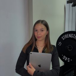 Studio Treningowe Martyna Poręba - Młoda kobieta w czarnej bluzce trzyma tablet na tle siłowni z obciążeniami. Profesjonalny wygląd, potencjalnie trenerka lub fizjoterapeutka prezentująca plan treningowy.