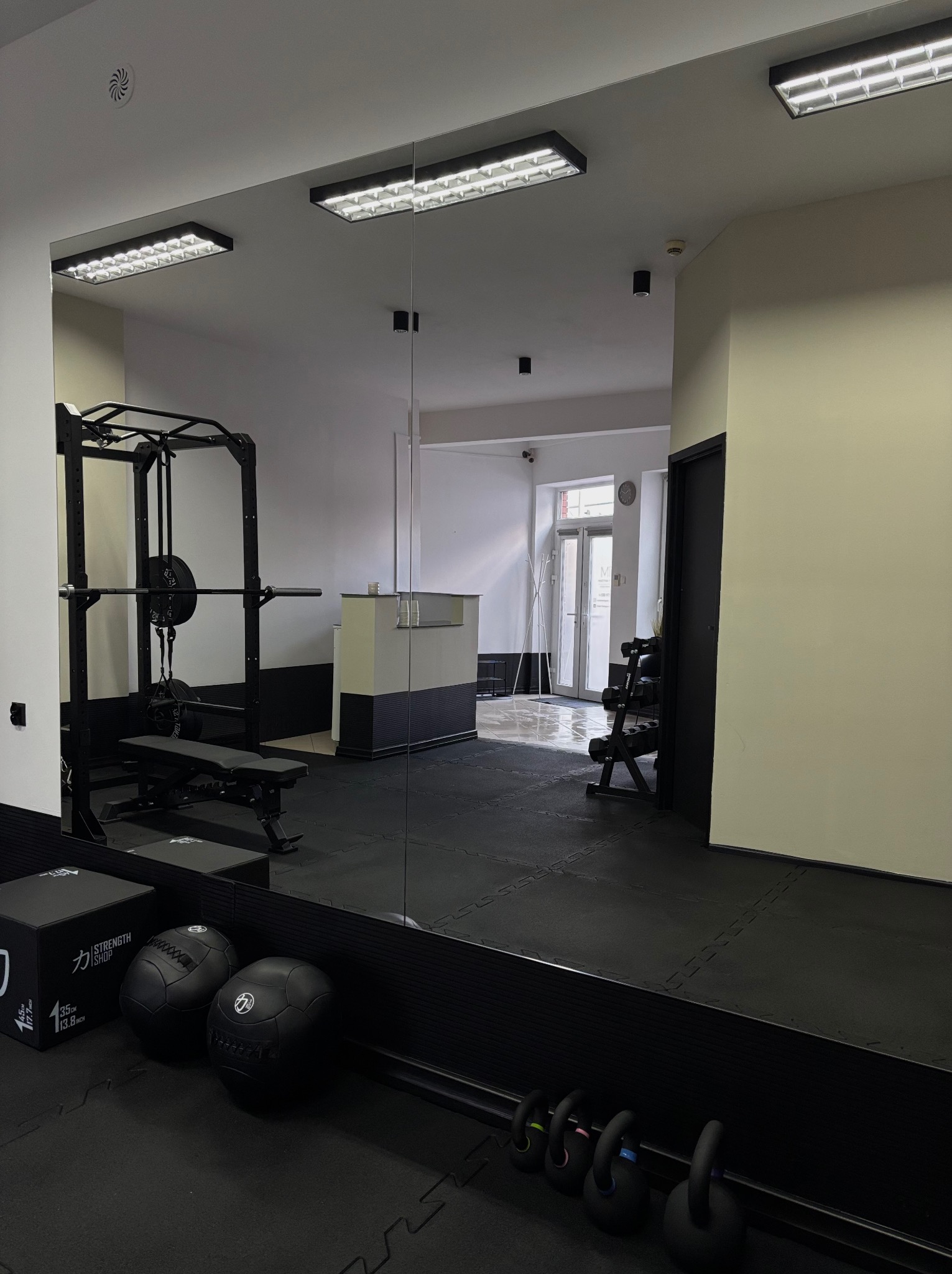 Widok sali treningowej z lustrem, ukazującym sprzęt do ćwiczeń: sztangę, kettlebells, piłki lekarskie. Gumowa podłoga i oświetlenie sufitowe.