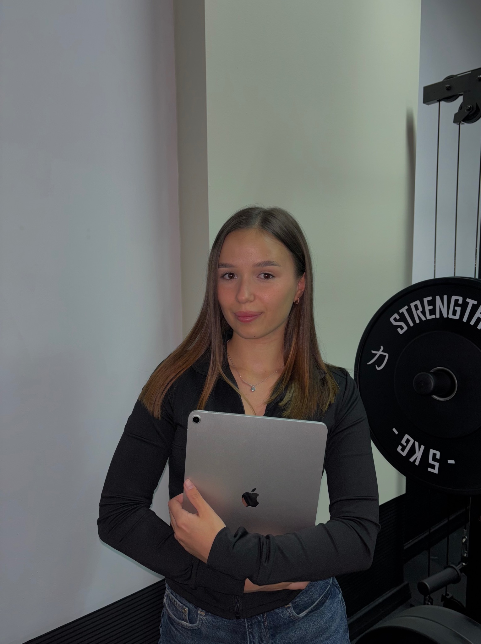 Młoda kobieta w czarnej bluzce trzyma tablet na tle siłowni z obciążeniami. Profesjonalny wygląd, potencjalnie trenerka lub fizjoterapeutka prezentująca plan treningowy.