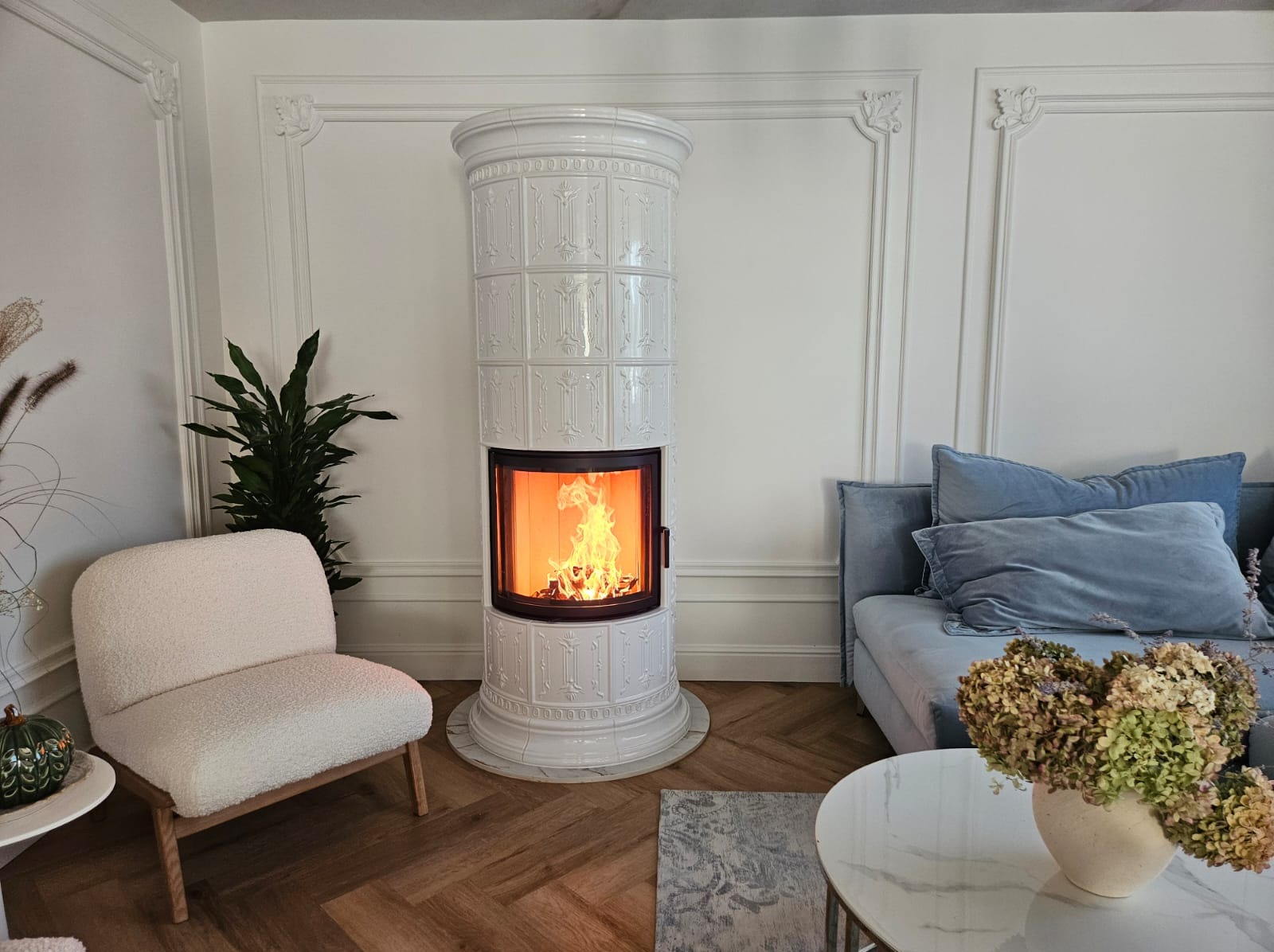 Biały, kaflowy piec kominkowy Schmid Rondo 55 51s w jasnym salonie z drewnianą podłogą w jodełkę i dekoracyjnymi listwami ściennymi. Płomienie wewnątrz.