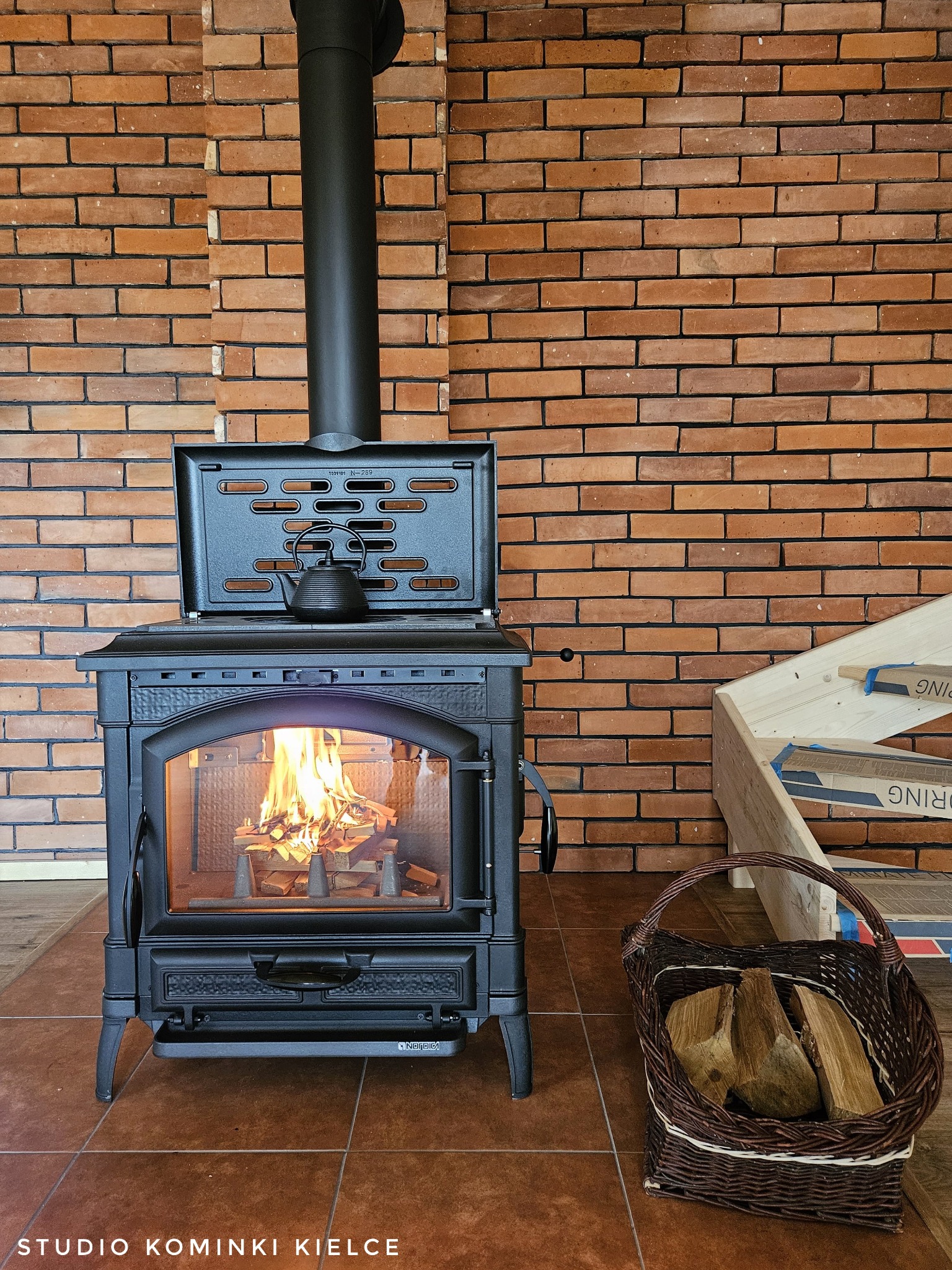 Żeliwny piec kominkowy La Nordica z paleniskiem, na którym płonie ogień. Na górze czajnik. Kosz z drewnem obok. Tło z cegły. Widoczne logo producenta.