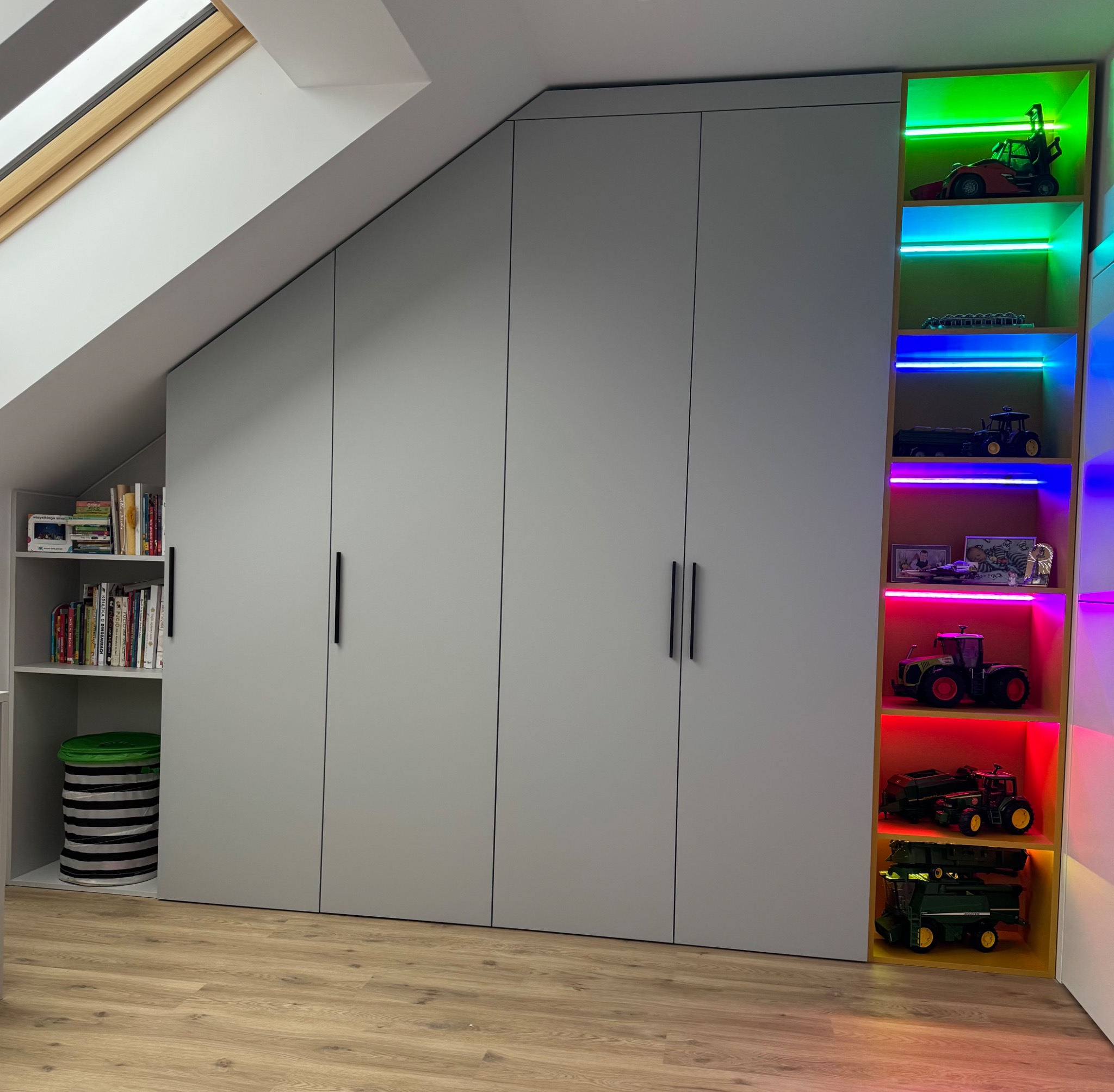Szara szafa wnękowa pod skosem dachu z otwartą półką na książki i podświetlaną półką z zabawkami, czarne uchwyty, jasna podłoga, okno dachowe.