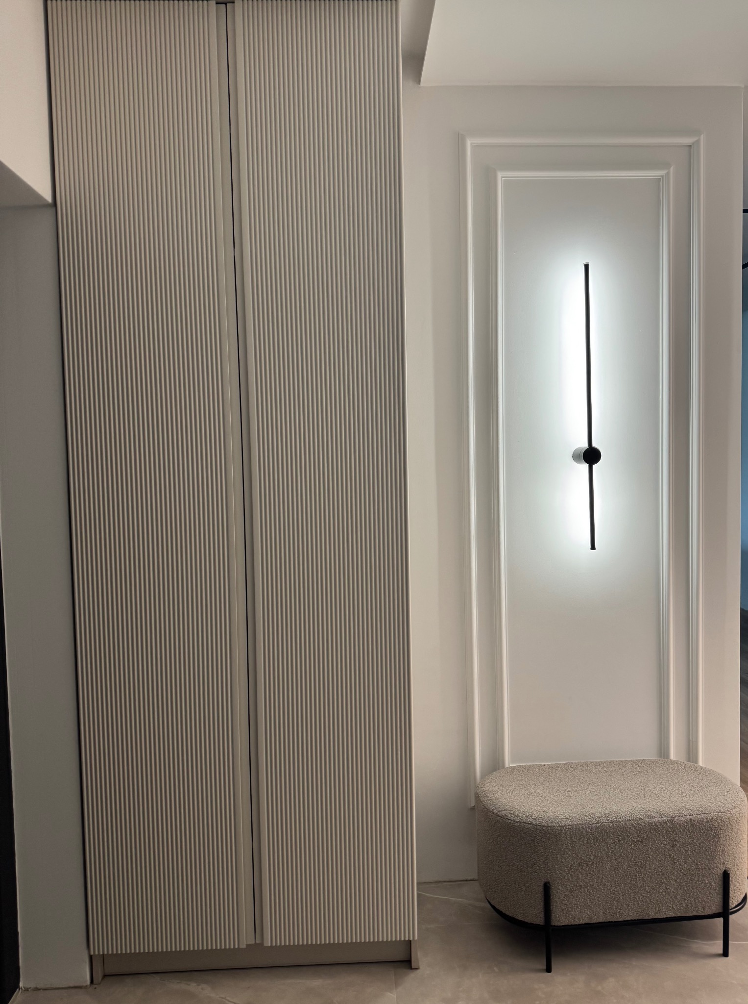 Nowoczesna szafa wnękowa z ryflowanymi drzwiami w odcieniach beżu, uzupełniona minimalistyczną lampą ścienną i pufą w neutralnej kolorystyce. Eleganckie wykończenie wnętrza.