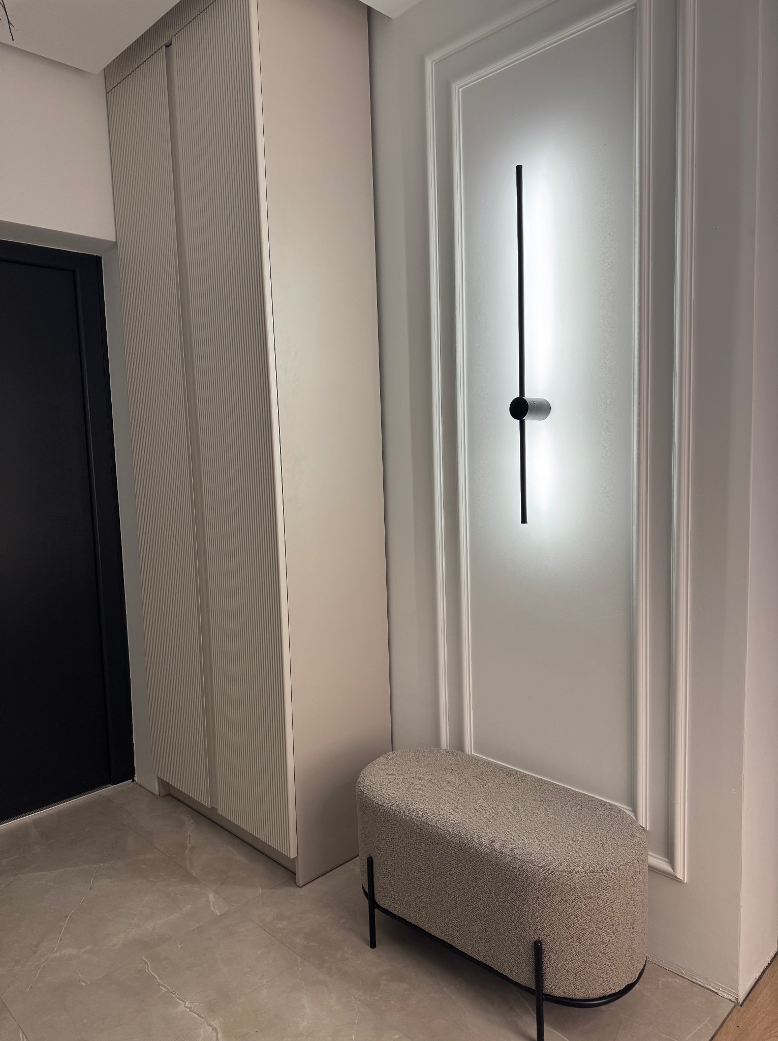 Nowoczesna szafa z ryflowanymi frontami w jasnym odcieniu, minimalistyczna lampa ścienna i tapicerowana ławka na marmurowej podłodze.