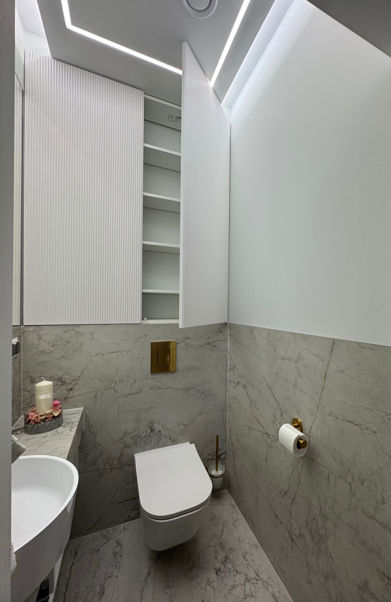 Nowoczesna toaleta z szafką na wymiar o ryflowanych frontach, marmurowymi płytkami i złotymi akcentami. Minimalistyczny design z oświetleniem LED.