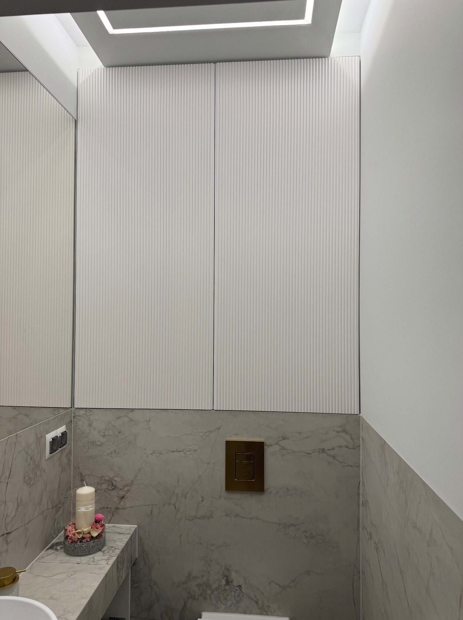 Białe, ryflowane szafki w łazience z marmurowymi płytkami i złotym przyciskiem spłuczki. Minimalistyczny design z oświetleniem LED.
