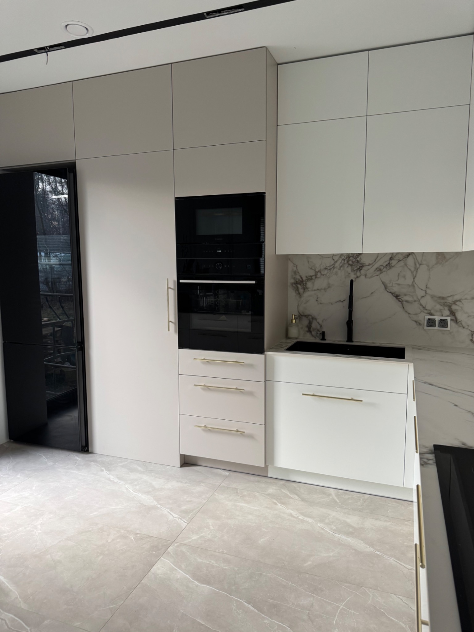 Nowoczesna, jasna kuchnia z zabudową na wymiar: białe fronty, złote uchwyty, marmurowy backsplash i czarny sprzęt AGD. Minimalistyczny design, eleganckie wykończenie.