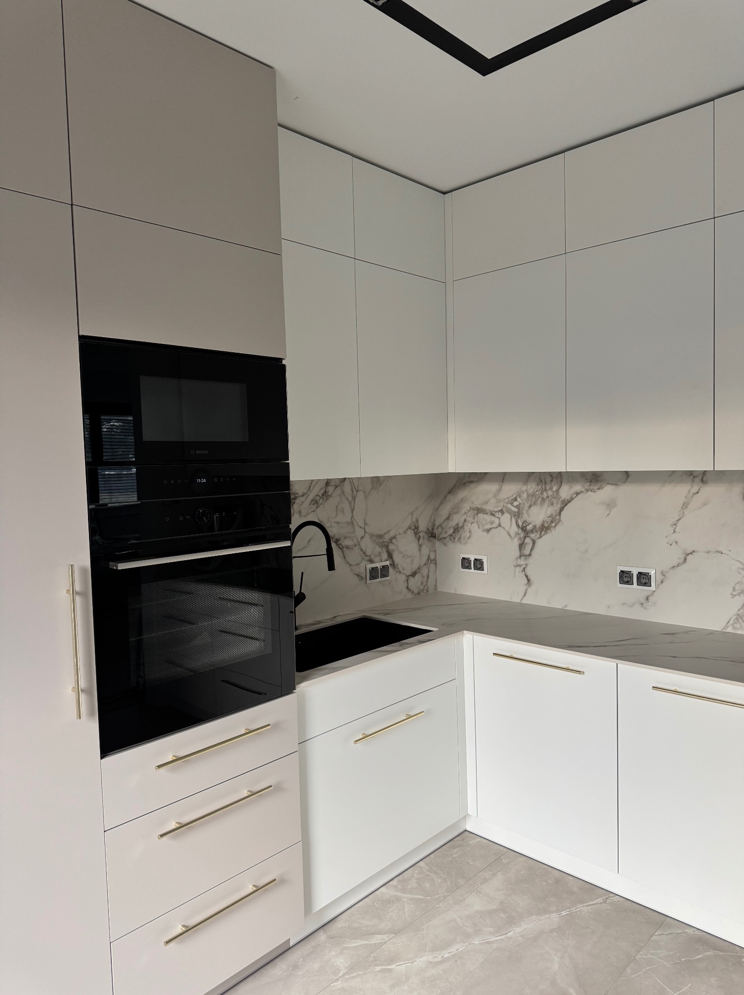 Nowoczesna, biała kuchnia z marmurowym blatem i złotymi uchwytami. Zabudowa z piekarnikiem i mikrofalówką. Minimalistyczny design z czarną armaturą i oświetleniem.