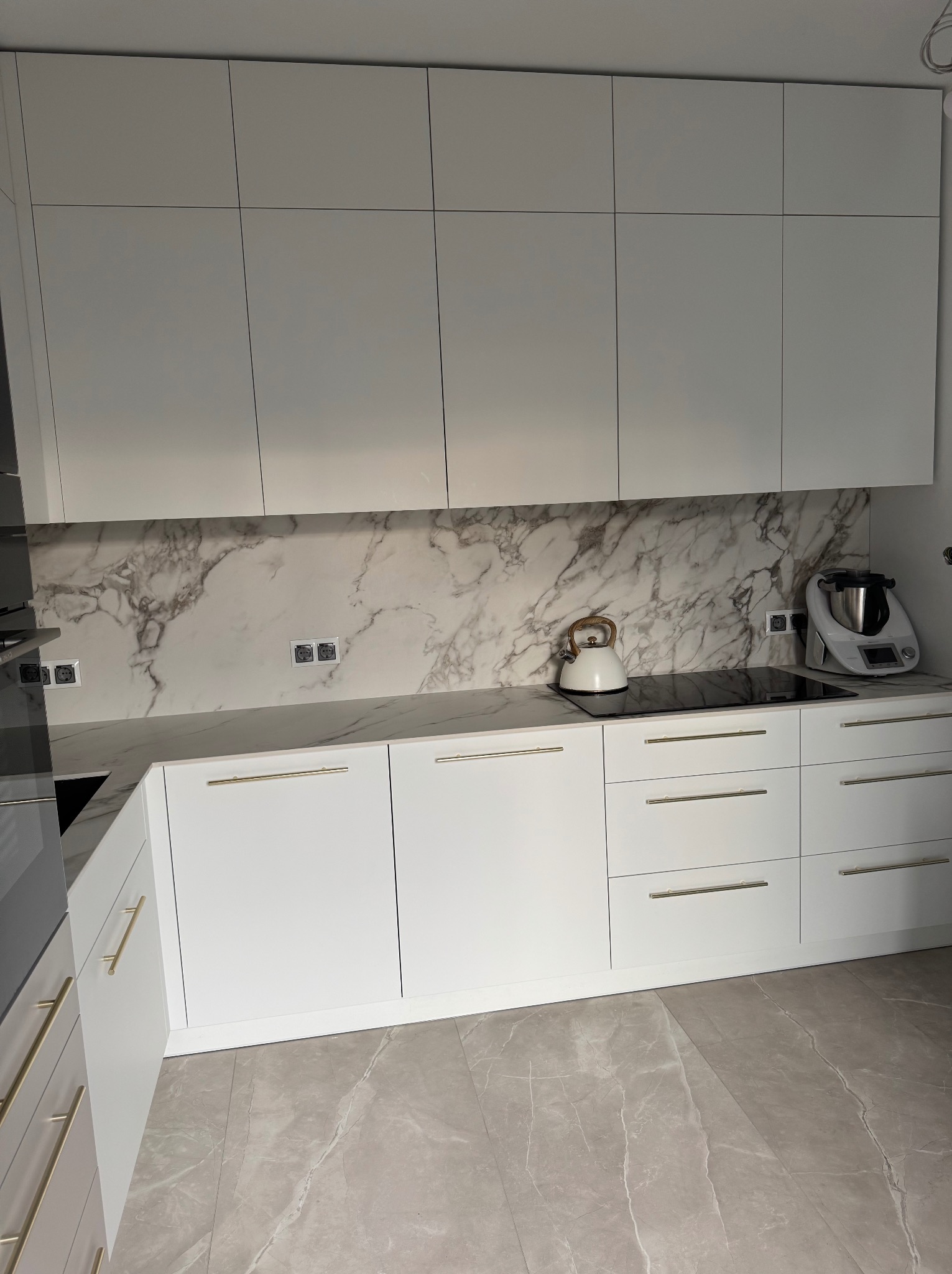 Biała kuchnia z marmurowym blatem i złotymi uchwytami, czajnik na płycie indukcyjnej, robot kuchenny. Minimalistyczny design, nowoczesne wykończenie.