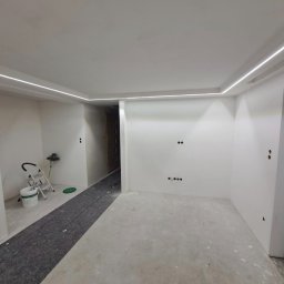 Kam-Sach-Wykończenia Wnętrz - Wnętrze po remoncie: białe ściany z subtelnym oświetleniem LED przy suficie, przygotowane do dalszych prac wykończeniowych, widoczne zabezpieczenia podłogi.