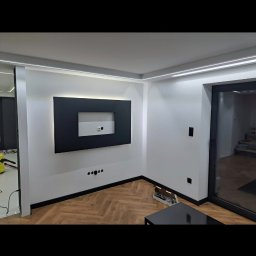 Kam-Sach-Wykończenia Wnętrz - Nowoczesny salon po remoncie: białe ściany, podłoga w jodełkę, czarna obudowa TV z podświetleniem LED i listwy przypodłogowe. Widoczne gniazdka elektryczne i włączniki.
