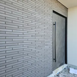 Elewacja budynku z szarej cegły klinkierowej i nowoczesnymi drzwiami wejściowymi z długim, czarnym uchwytem. Widoczne drobne prace wykończeniowe przy fundamencie.