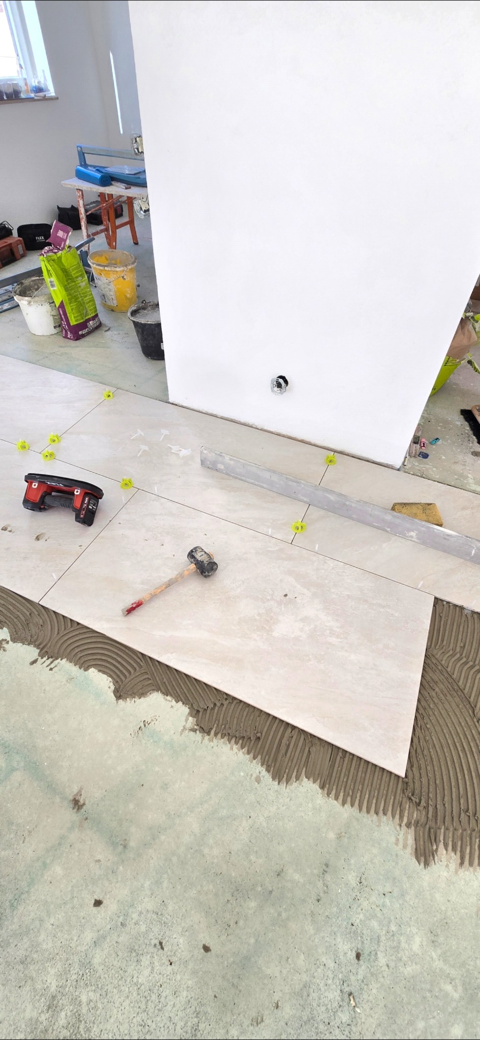 Układanie jasnych płytek podłogowych na klej, widoczne narzędzia: poziomica, gumowy młotek. Żółte krzyżyki dystansowe. Praca w toku, wykończenie podłogi.