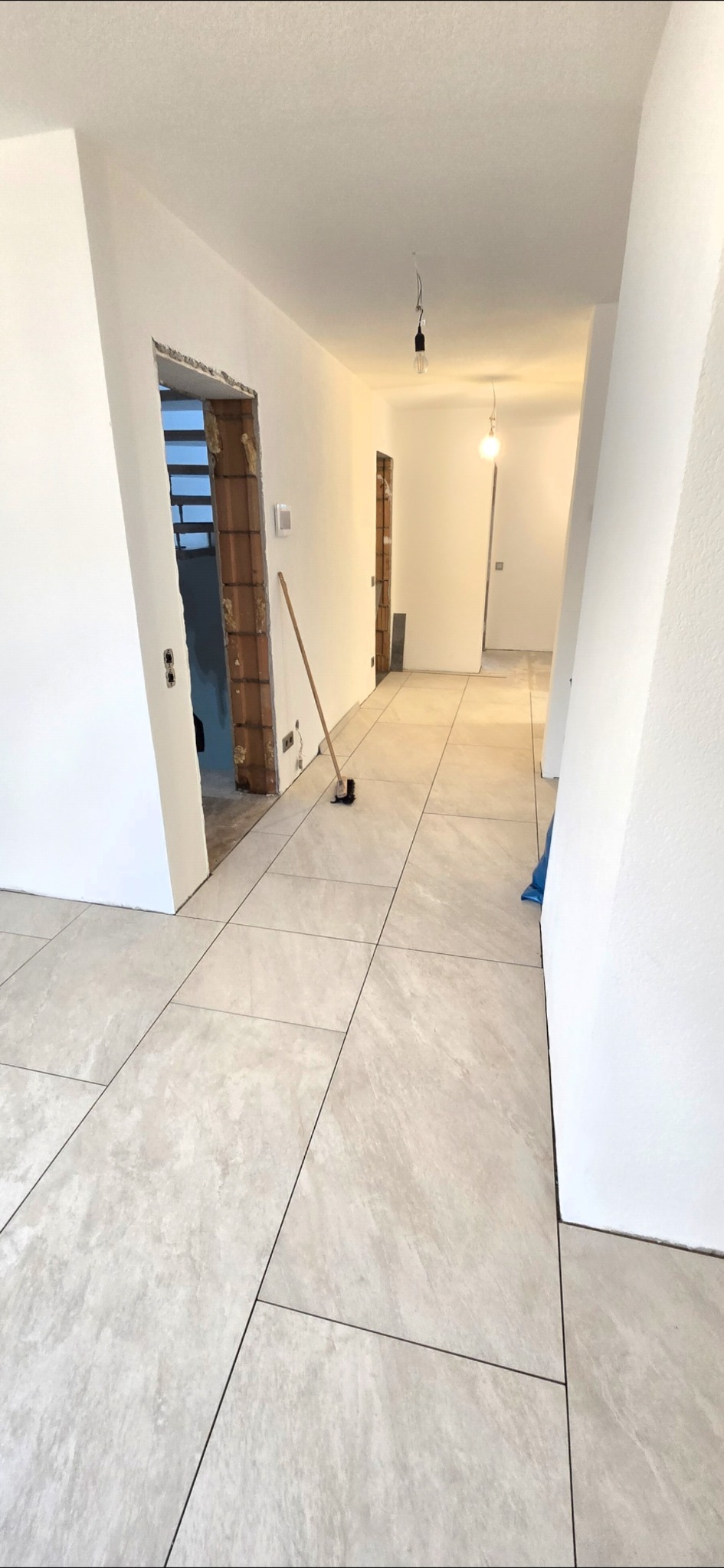 Jasne, duże płytki podłogowe w korytarzu w trakcie remontu. Widoczne przygotowane ościeżnice i surowy stan ścian. Na podłodze leży miotła.