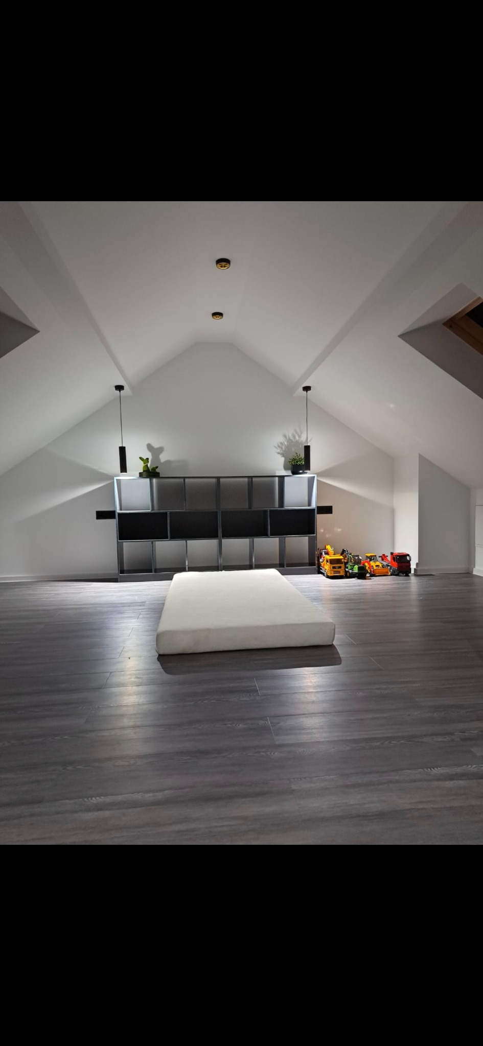 Poddasze po remoncie: szara podłoga, białe ściany i sufit, czarne lampy wiszące. Regał z dekoracjami i zabawki w tle. Na podłodze leży materac.