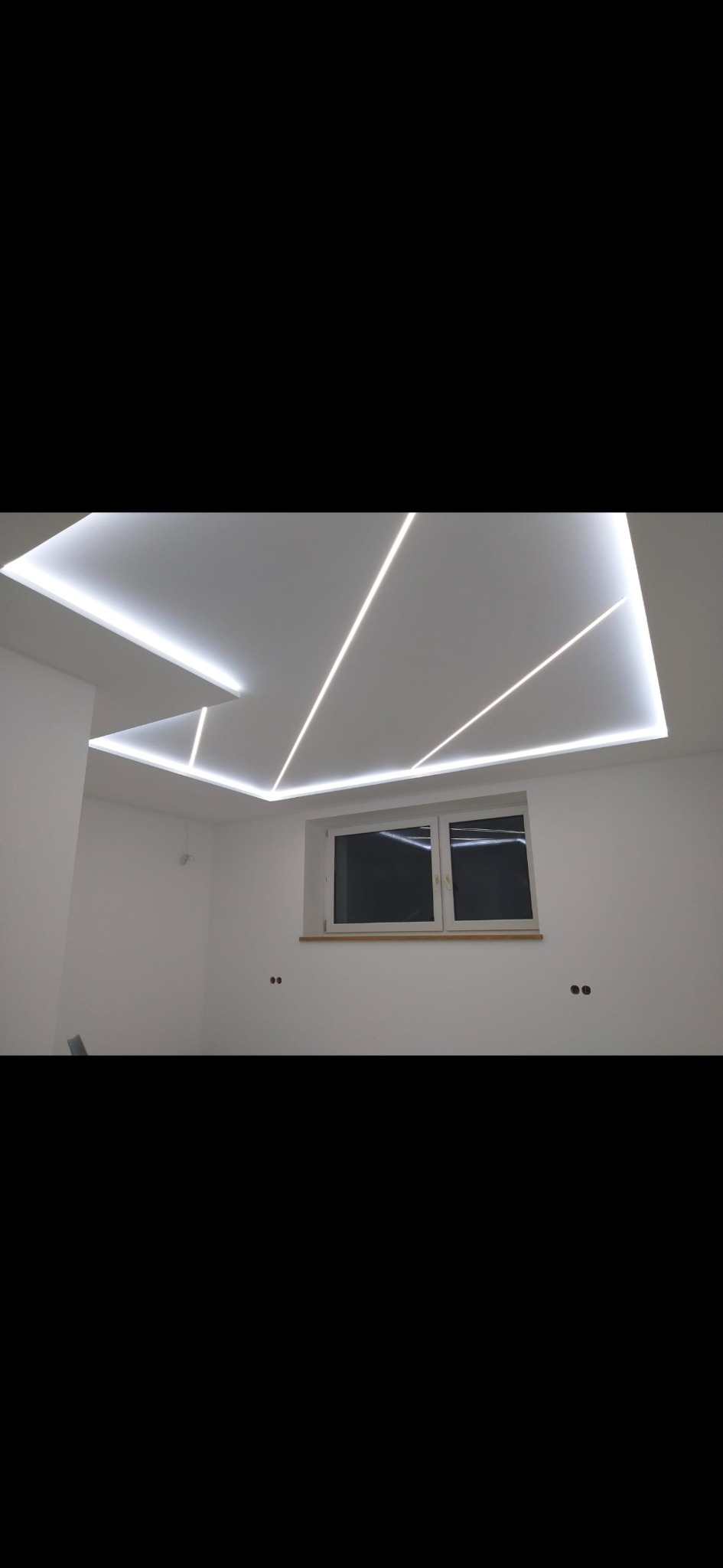 Białe wnętrze pokoju z oknem i designerskim sufitem podwieszanym z liniowym oświetleniem LED tworzącym geometryczne wzory.
